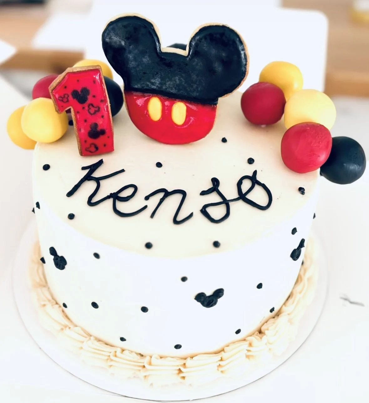 Pastel de cumpleaños con decoraciones de Mickey Mouse, un símbolo de Mickey en colores rojo, negro y amarillo, y una vela en forma de cifra uno con corazones y dibujos de Mickey Mouse. La palabra 'Kensho' escrita en glaseado negro en la parte superior del pastel.