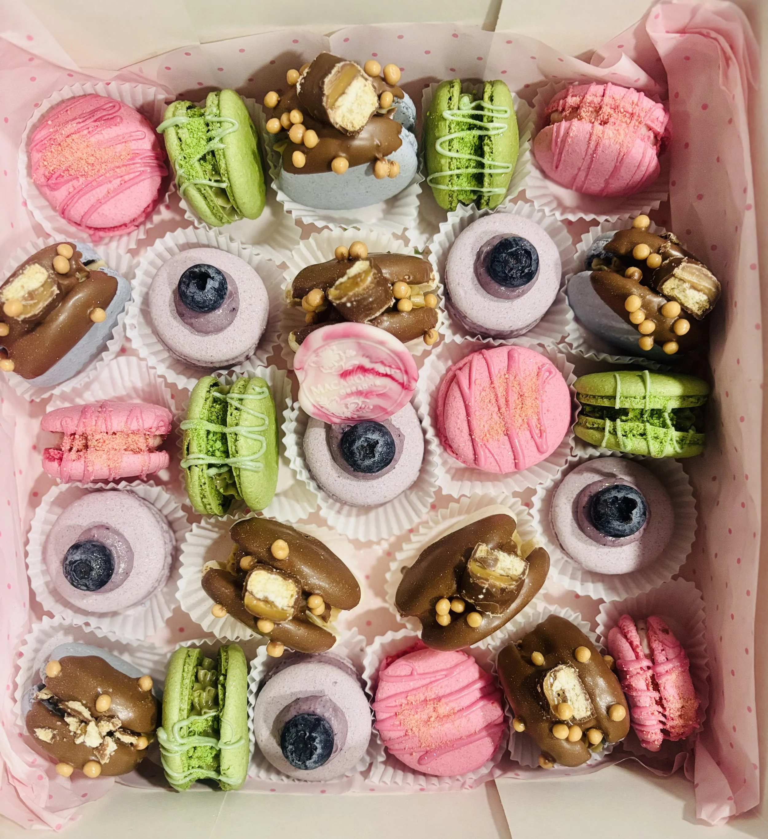 Caja de macarons decorativos en diferentes colores y sabores, con decoraciones de chocolate, frutas y toppings variados.