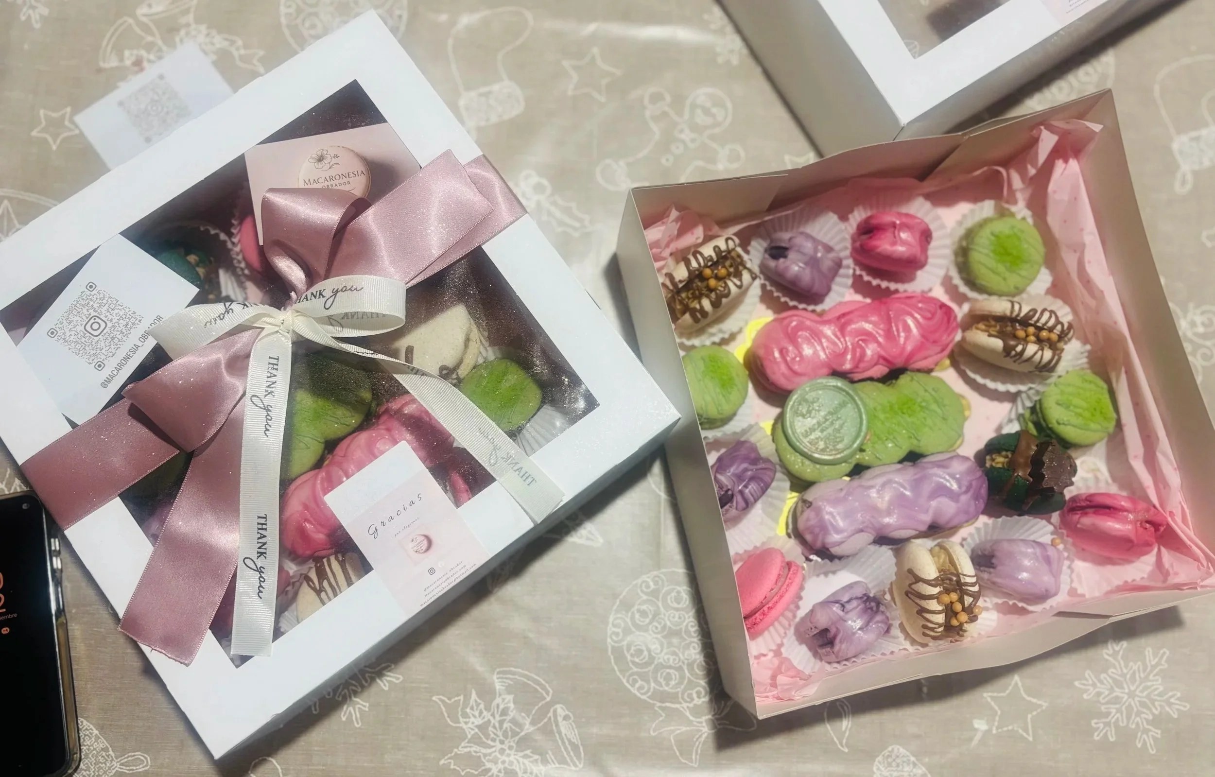 Caja de regalo con galletas decoradas de colores rosa, verde, morado, y beige, algunas con decoración de chocolate, en una bandeja forrada con papel rosa, y otra caja decorada con lazo rosa y una etiqueta que dice 'Gracias'.