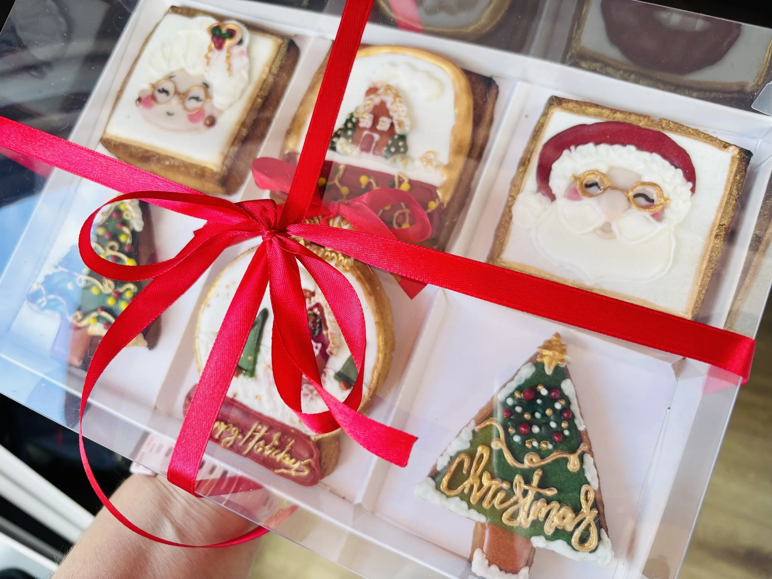 Caja de galletas decoradas de Navidad, con motivos como Santa Claus, árbol de Navidad, reno y nieve, adornadas con glaseado y colores festivos, atadas con cinta roja.