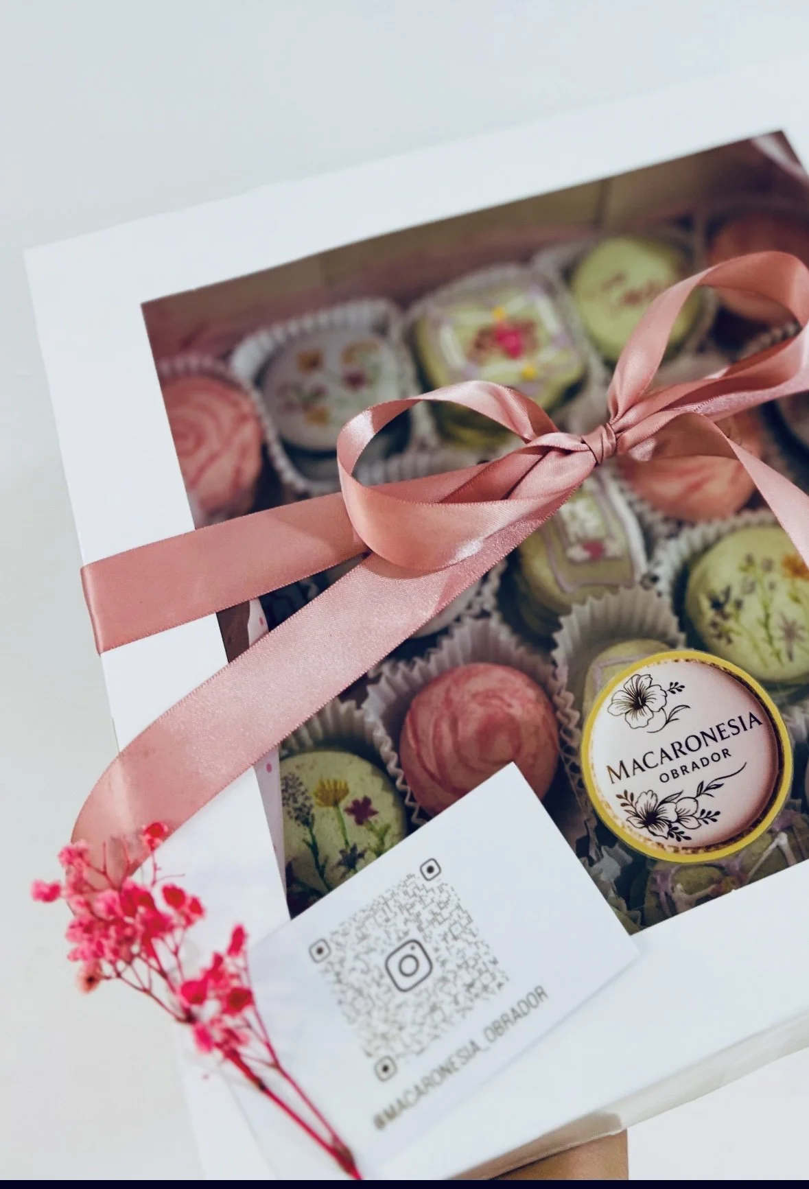 Caja de chocolates artesanales decorados con ilustraciones florales, envueltos en una caja blanca con ventana y una cinta de satén rosa.