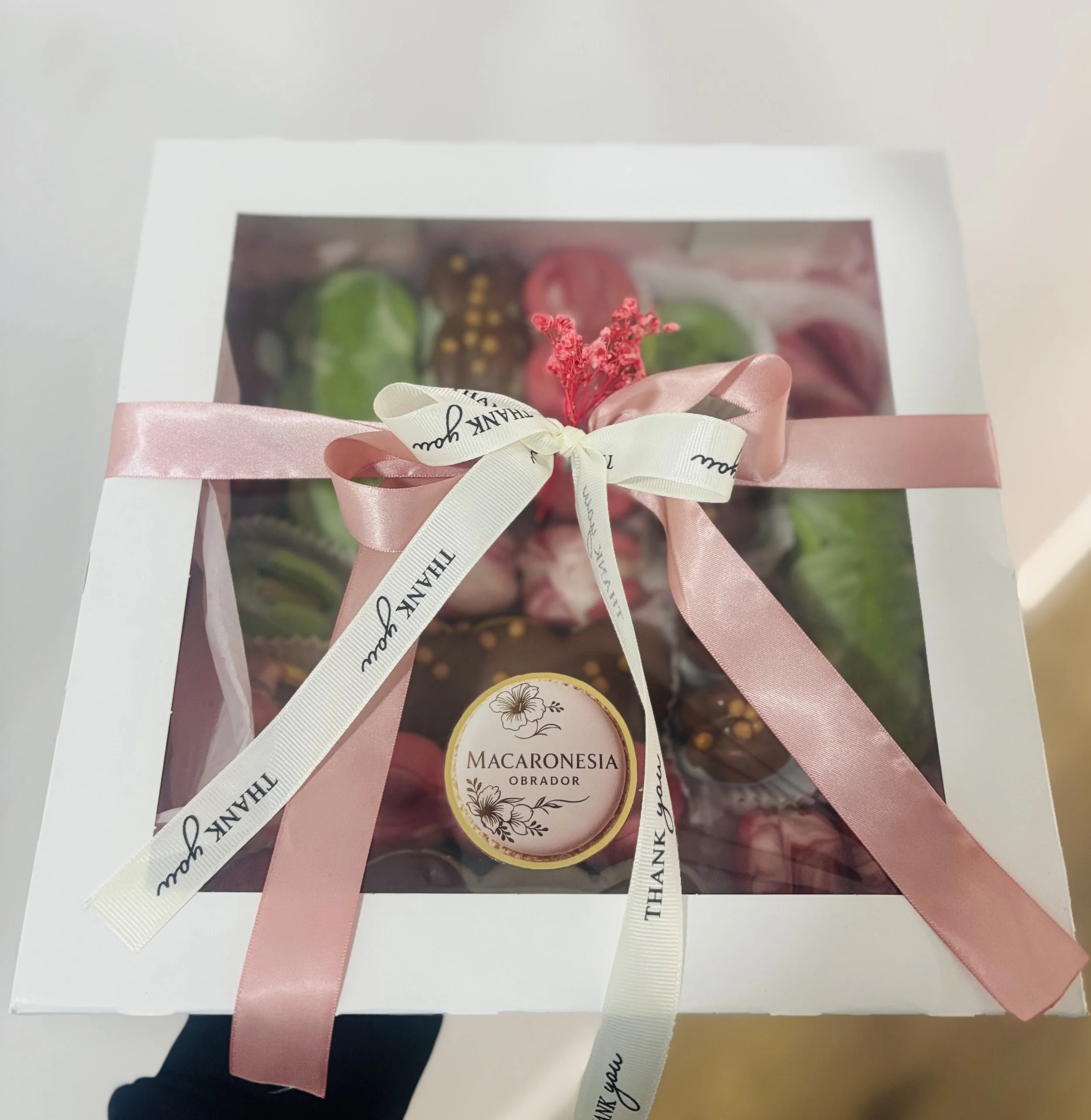 Caja de chocolates decorada con la etiqueta 'Macaronesia Obrador', adornada con cinta rosa y blanca, y flores rosadas en la parte superior, vista desde arriba.