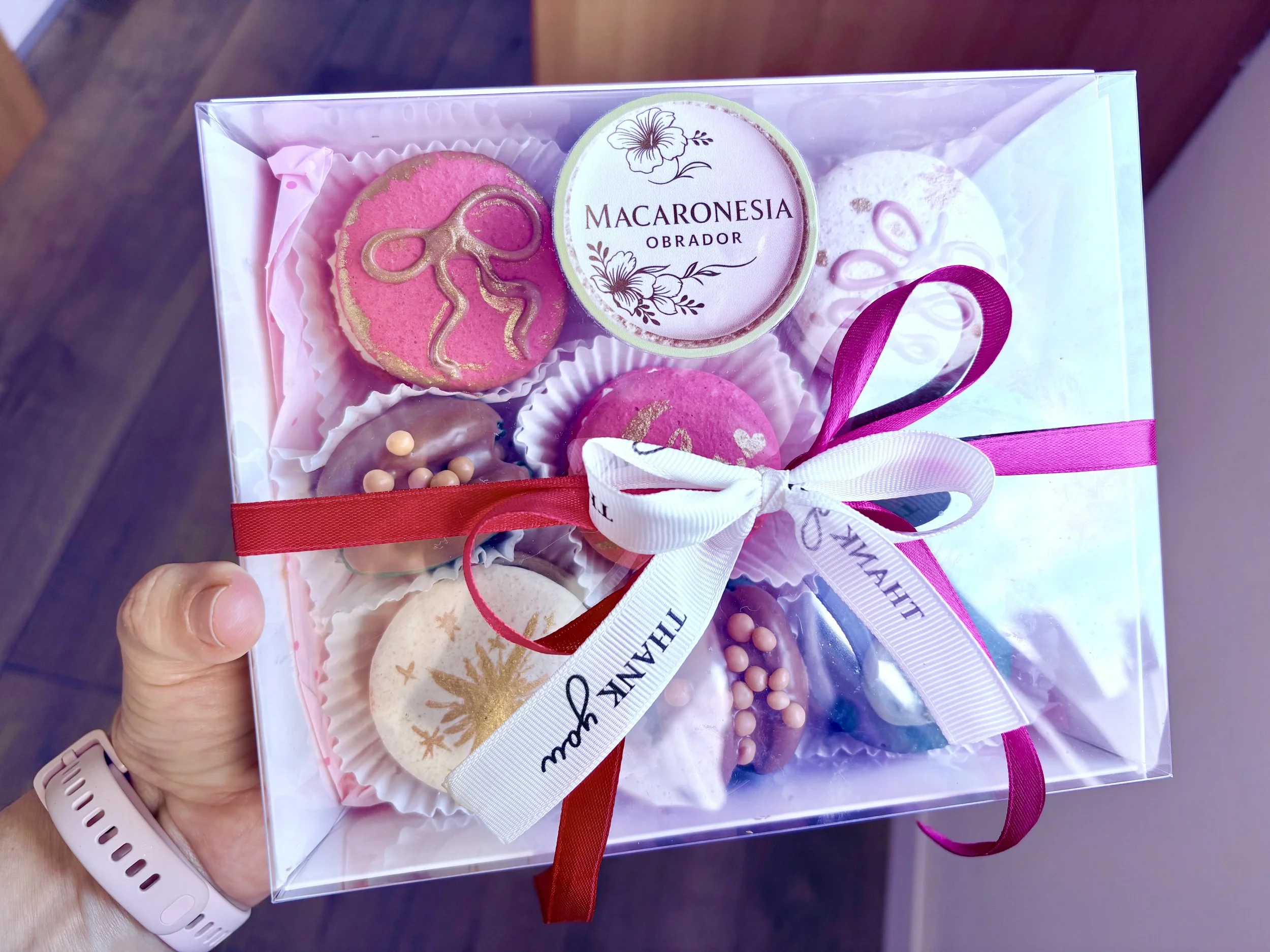 Caja con varios macarons decorados en tonos rosas, blancos y marrones, con la etiqueta 'Macaronesia Obrador' en la parte superior, adornada con un lazo blanco y uno rosa.