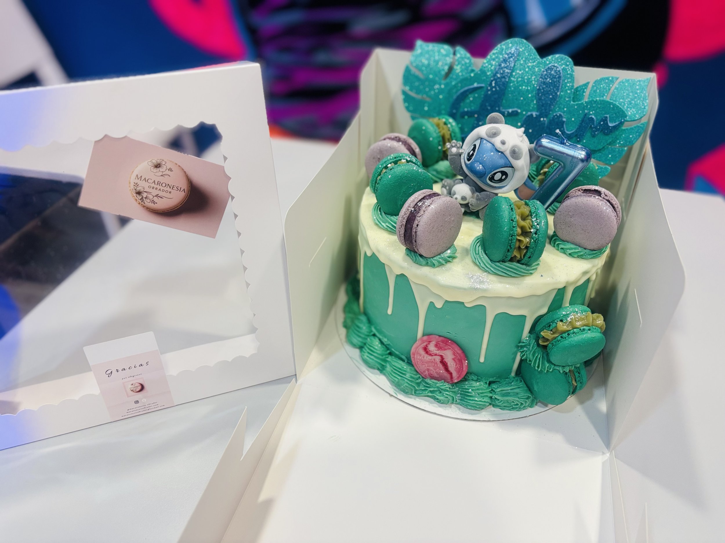 Pastel de cumpleaños con decoración en tonos verdes y lilas, con macarons en la parte superior, figura de un robot azul y una hoja decorativa, en caja blanca sobre mesa