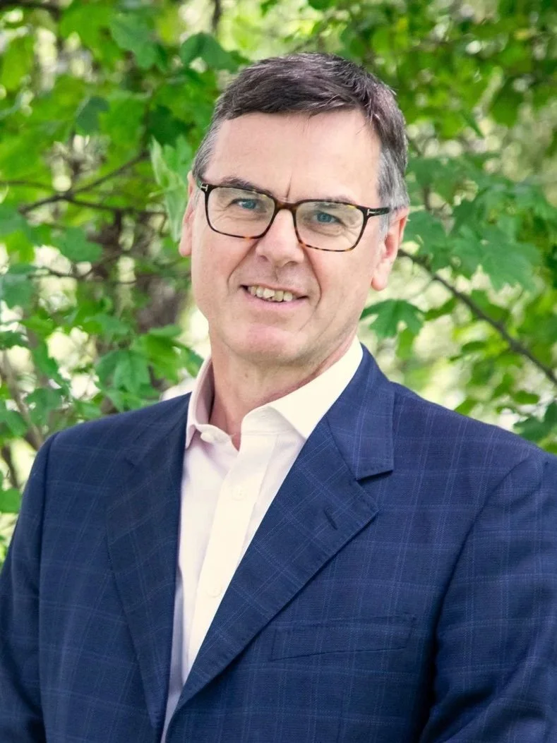 A middle-aged man with short dark hair, glasses, wearing a dark blue suit and a white shirt, standing outdoors in front of green foliage.