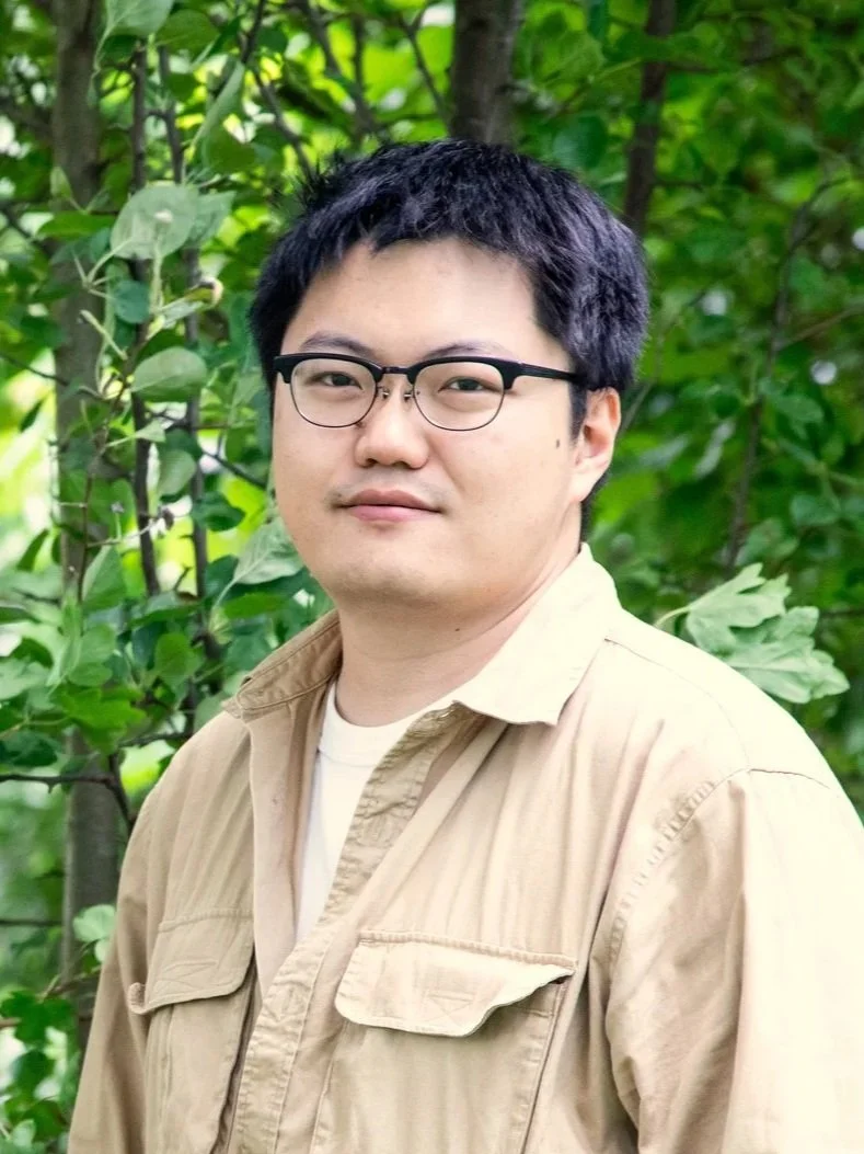 A young man with glasses and short black hair stands outdoors in front of green foliage, wearing a beige jacket and light-colored shirt.