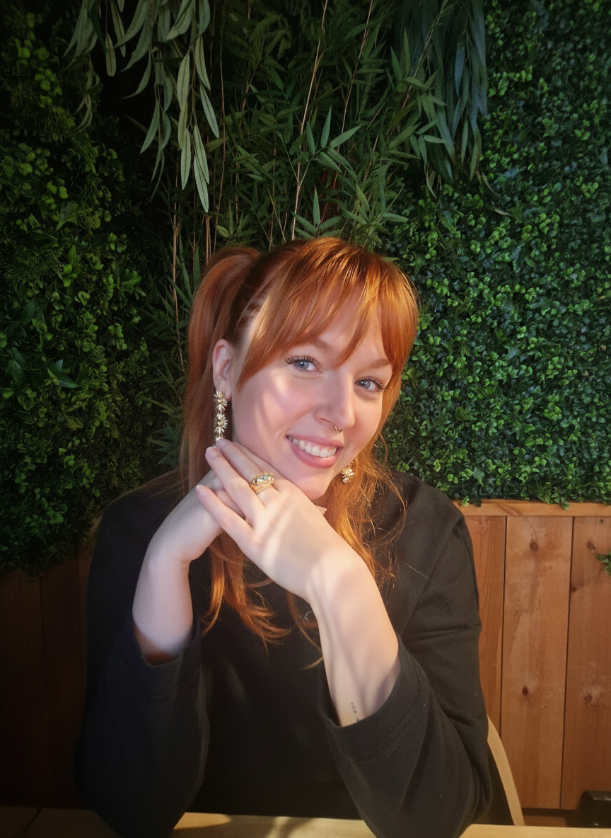 Vrouw met rood haar en lichte huid die glimlacht, zittend bij een tafel met een groene groene muur op de achtergrond. Yaihara van der Welle, Home of YAURA, Eigenaresse, Energetisch therapeut, Energetische therapie in Arnhem en ervaringsdeskundige.