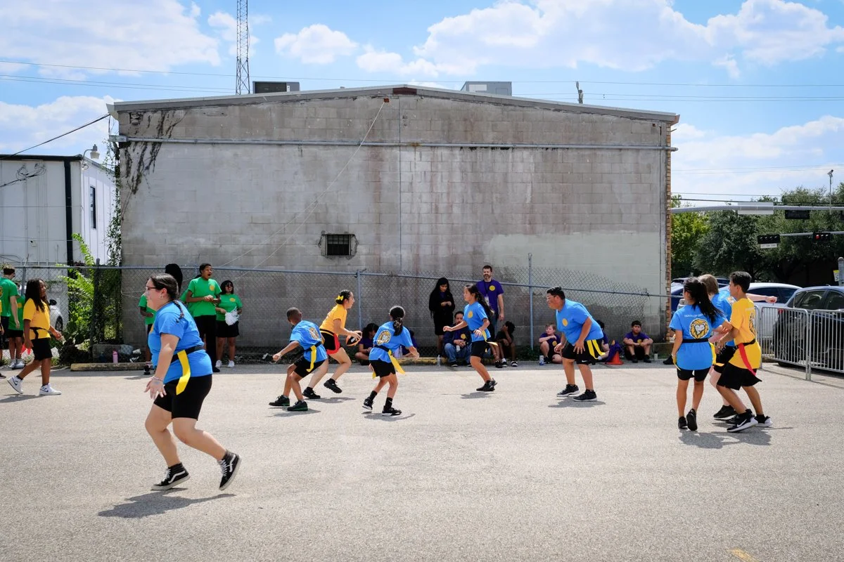 Children playing a game outdoors, with some wearing blue and others wearing yellow shirts, while adults watch from the side and some sit on benches, against an urban background with a large concrete wall and a fence.