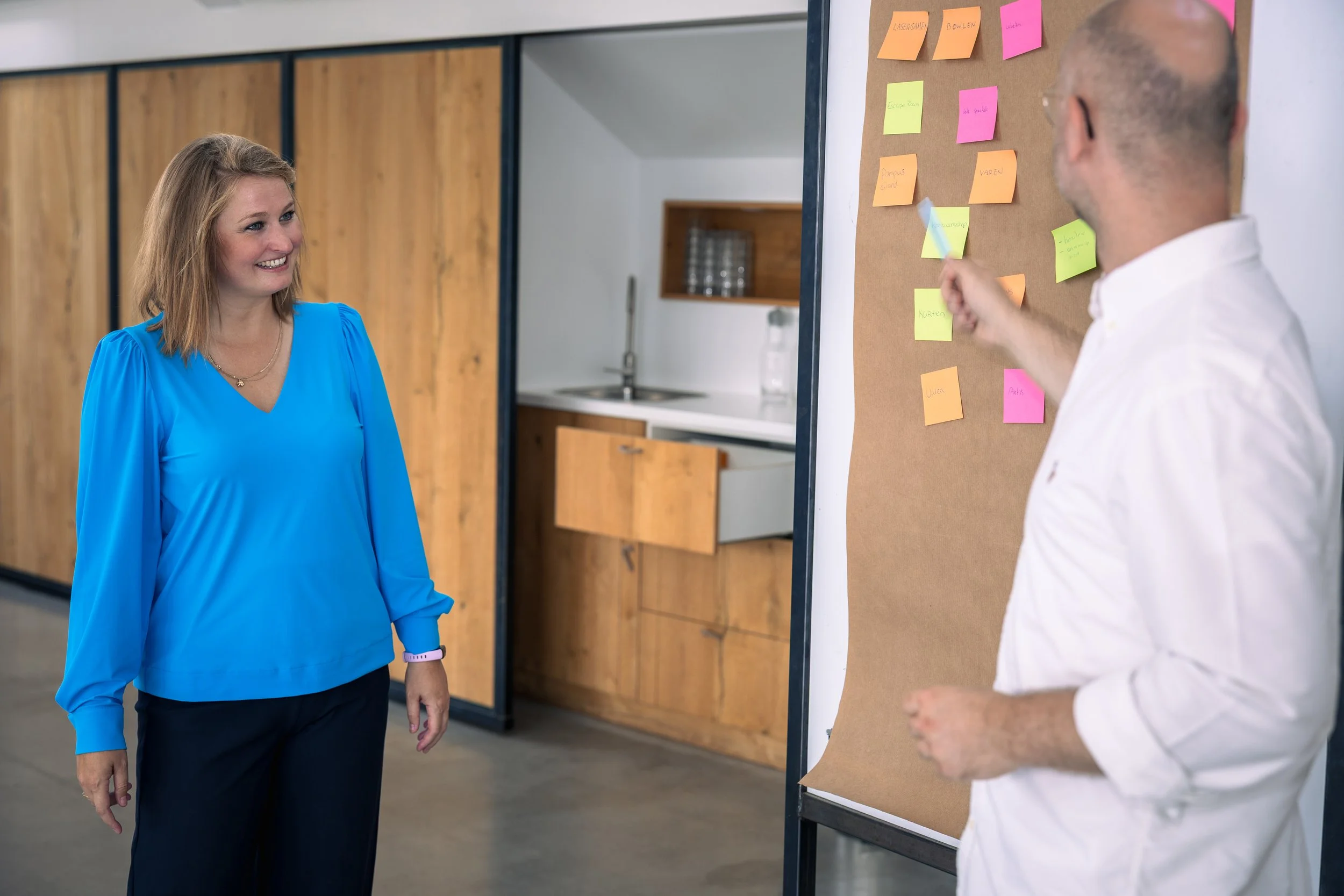 Een vrouw met een blauwe blouse en een man met een witte overhemd praten in een kantoorruimte. De man wijst naar post-it notities op een kartonnen prikbord.