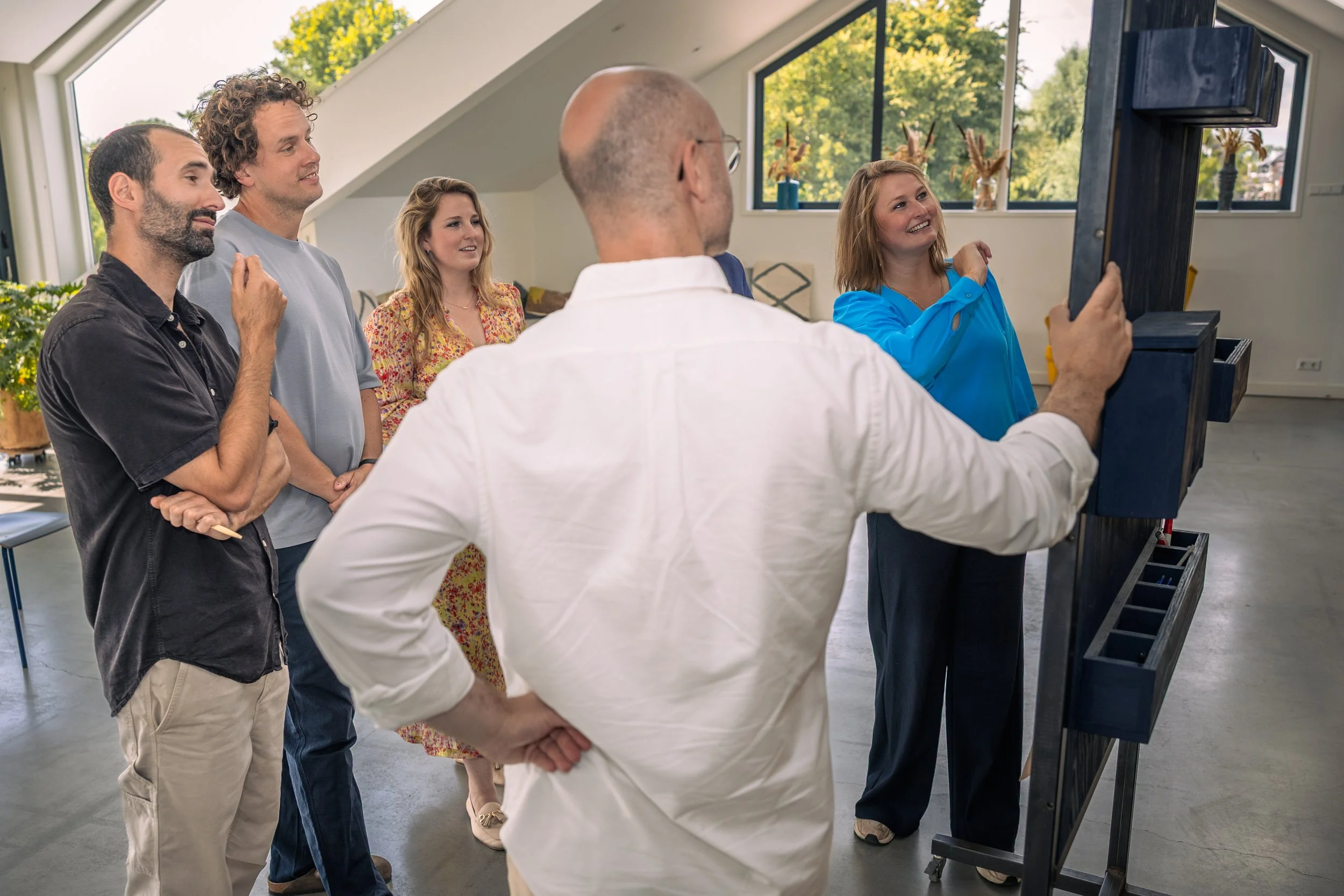 Een groep mensen staat in een kamer met grote ramen, terwijl een vrouw in een blauwe blouse een presentatie geeft met een schoolbord. De mensen kijken aandachtig, de sfeer lijkt informatief en ontspannen.