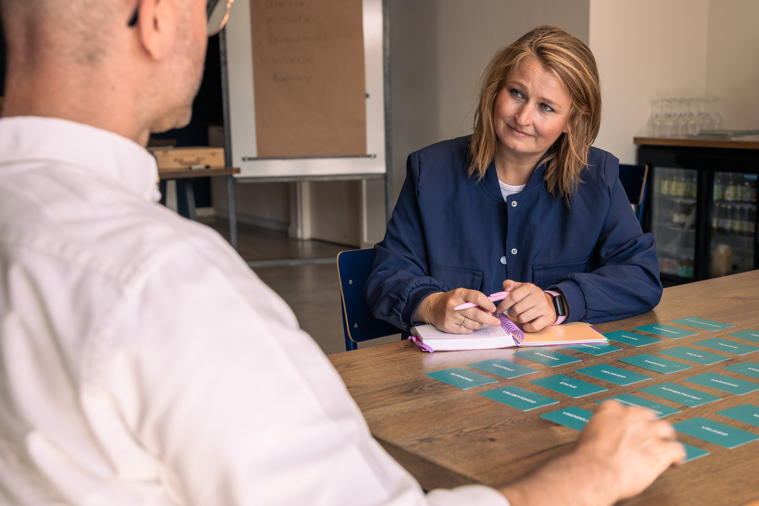 Vrouw zit aan tafel met kaarten voor een gesprek met een man, notitieboek en pen, in een vergaderruimte.