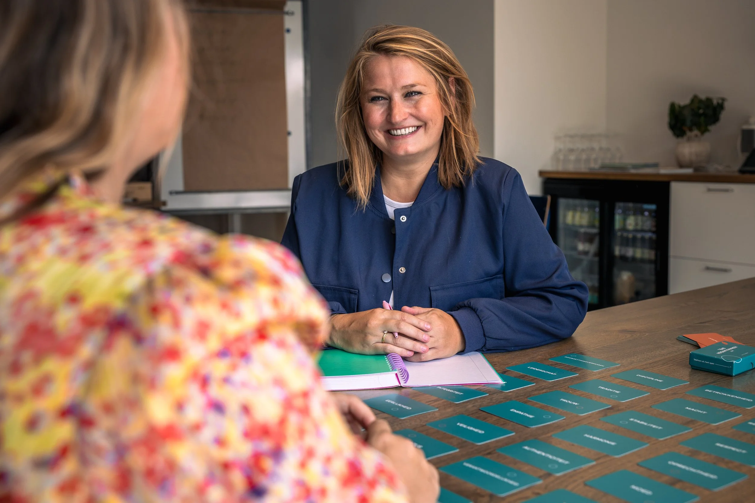 Floor Waterreus coaching, 1 op 1 coaching, persoonlijk leiderschap. Twee vrouwen zitten aan een tafel, de ene vrouw lachend, de andere vrouw gedeeltelijk zichtbaar. Op tafel liggen kaarten en een notitieboekje.