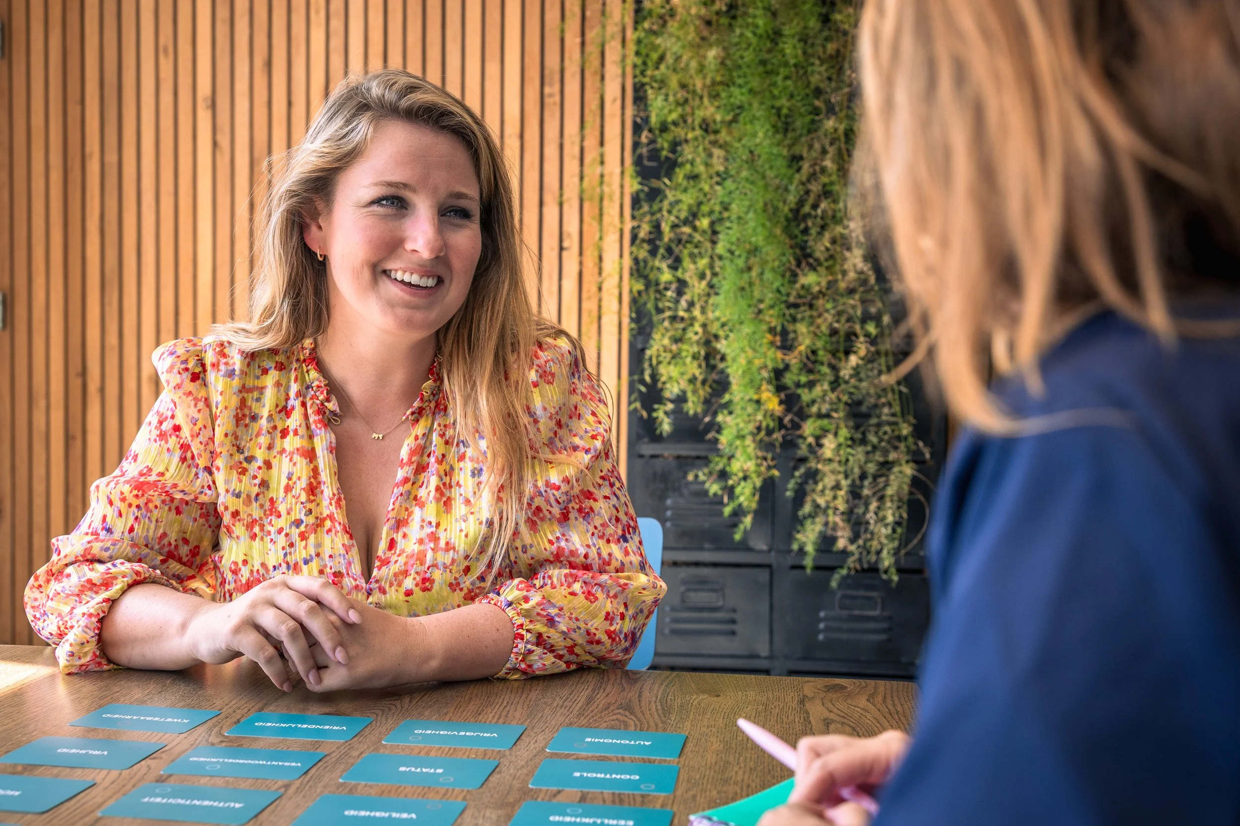 Vrouw die in een gesprek zit, lachend, aan een tafel met blauwe kaartjes en een pen.