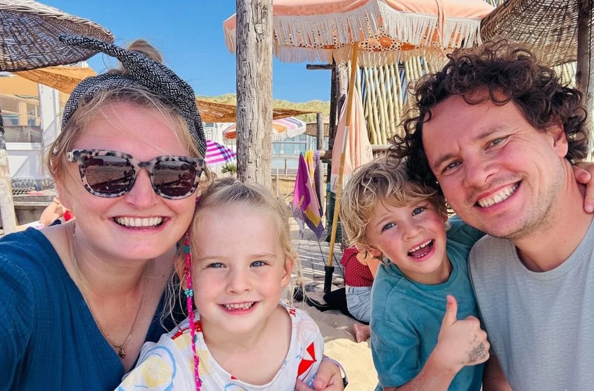 Een gezin op het strand onder een parasol, iedereen lacht en poseert voor de camera.
