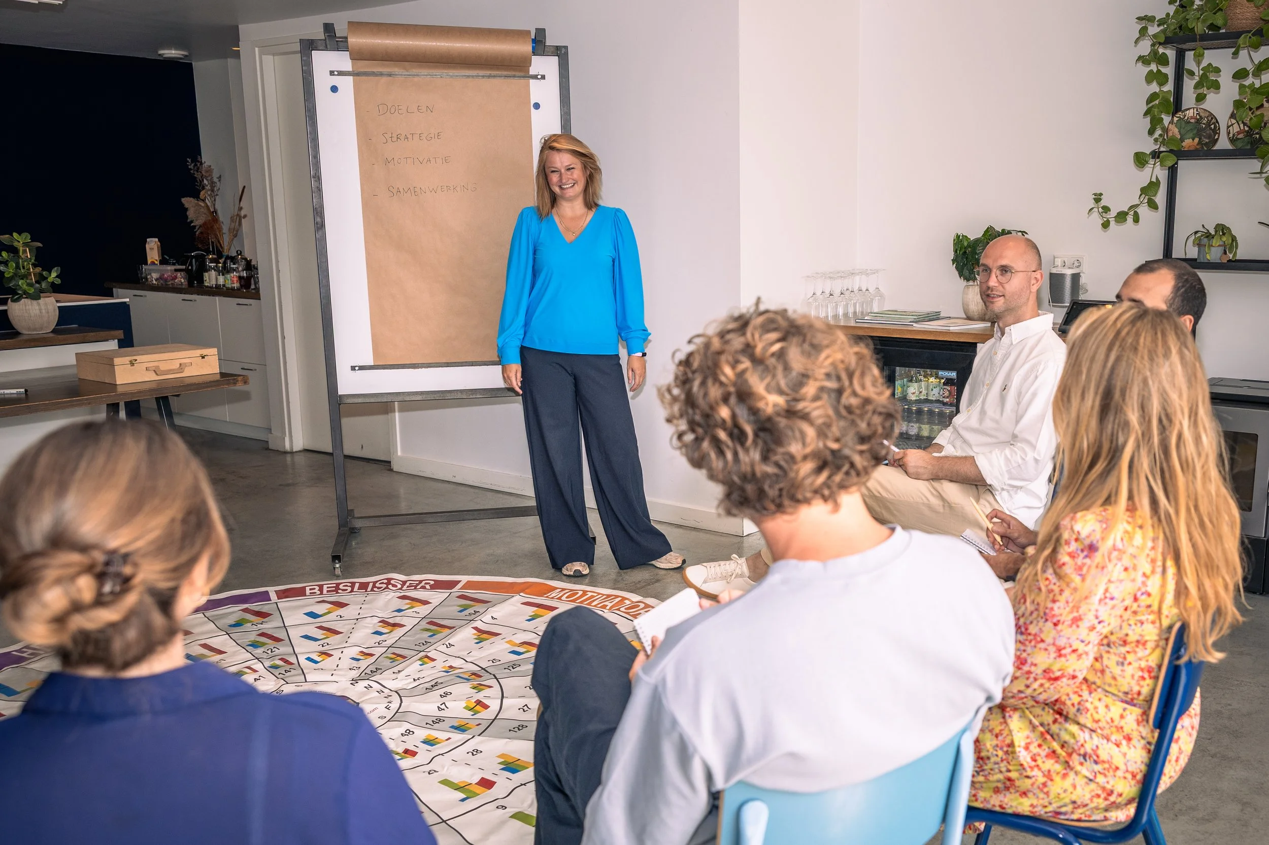 Een vrouw staat voor een groep mensen die een presentatie bijwonen in een modern kantoor. Ze lachen en er is een flipover met woorden als "Doelen", "Strategie", "Motivatie" en "Samenwerking". Mensen zitten op stoelen, sommigen maken notities. Op de vloer ligt een kleurrijk bord of spel.