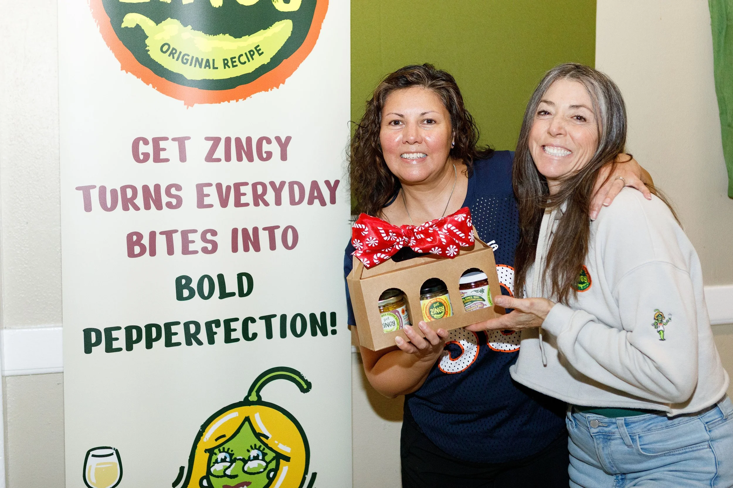 Smiling women, embracing closely, holding a Get Zingy Zing Pack variety of jars next to a sign that highlights Get Zingy's bold pepperfection.