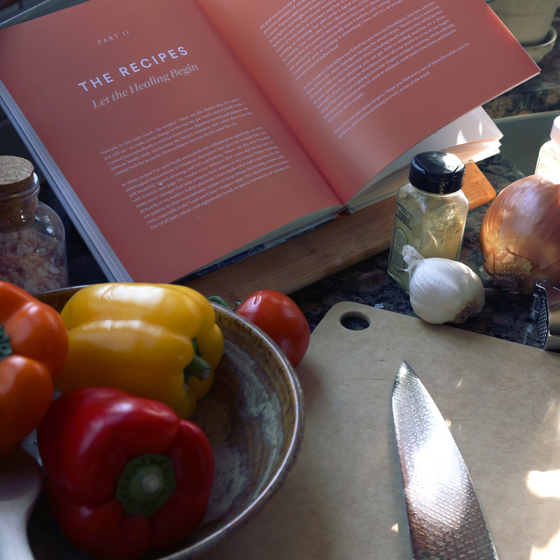 A counter full of ingredients with a recipe that highlights peppers, salt, garlic, tomatoes, a cutting board, knife and ceramic bowl.