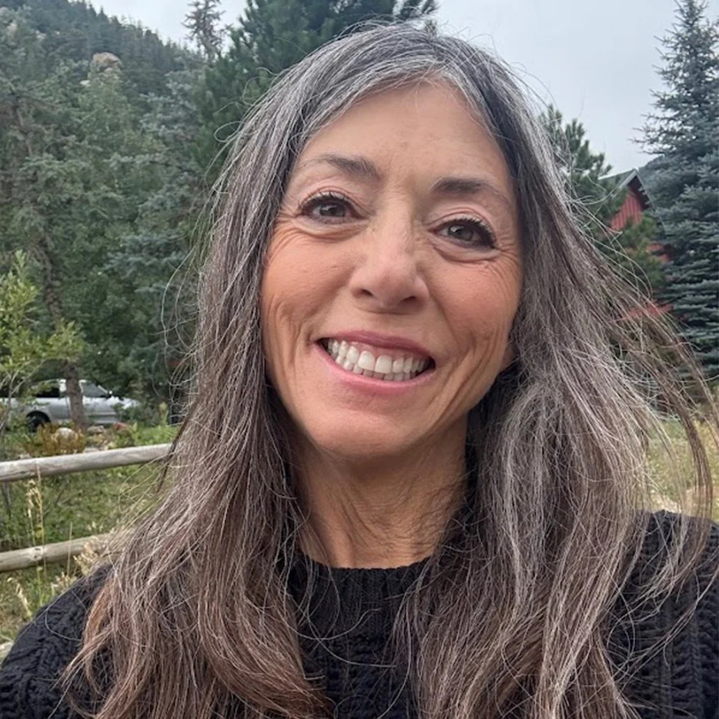A woman with long gray hair smiling outdoors, with trees and a fence in the background. She is the owner of Get Zingy pepper relish.
