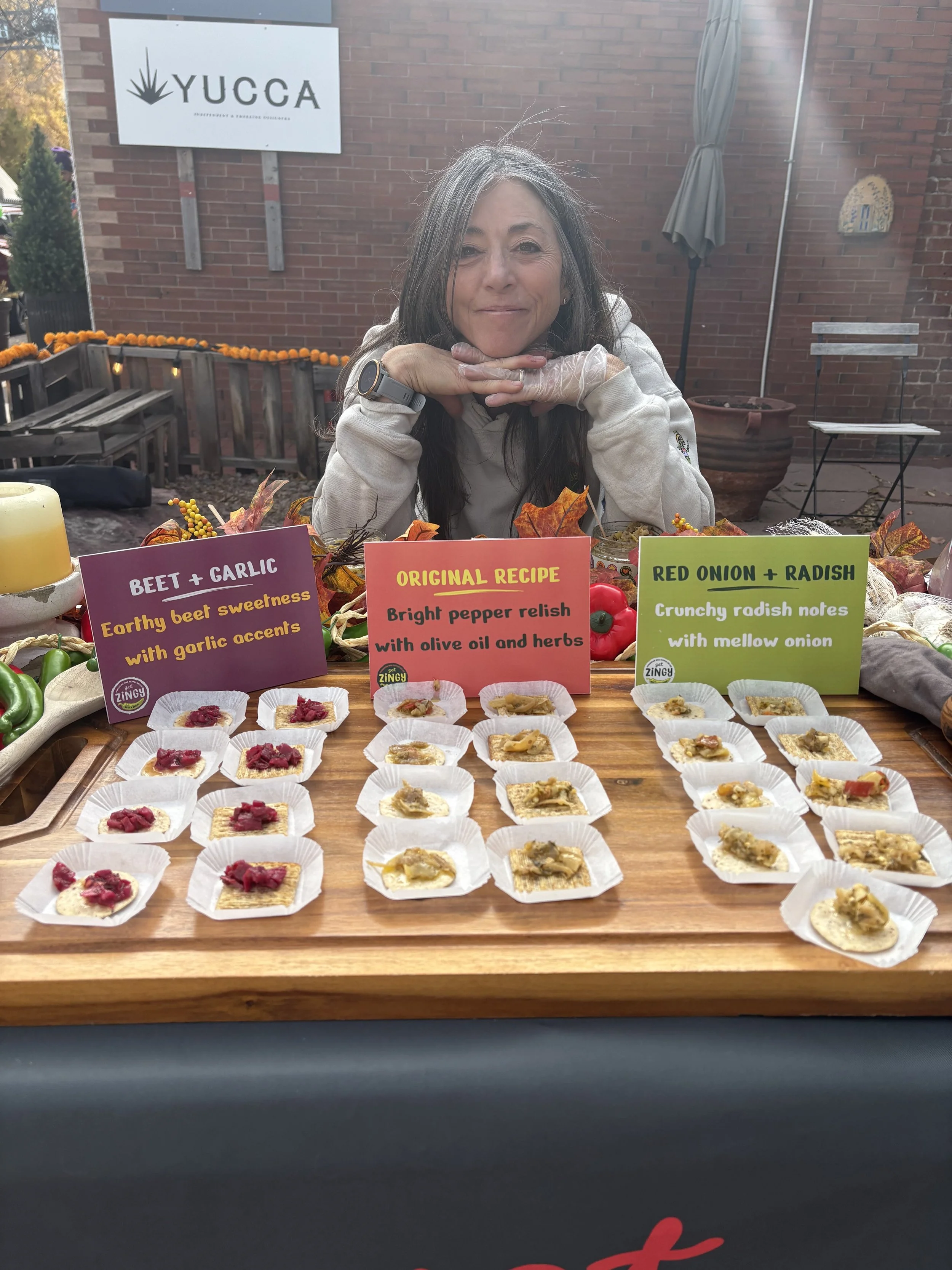 A woman sitting behind a table with sample food tastings labeled with signs: 'Beet + Garlic,' 'Original Recipe,' and 'Red Onion + Radish.' The table features small plates of food topped with different spreads or toppings, and the woman is smiling wit