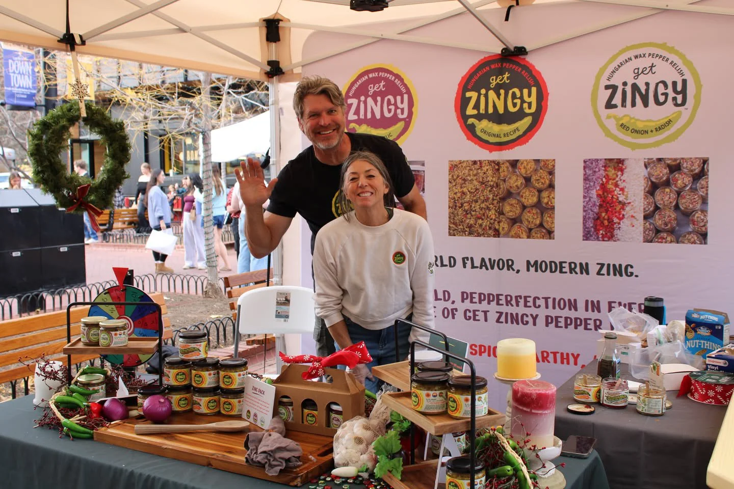 Two people standing behind a table at a market stall, smiling and waving. The stall has jars of a product called Get Zingy, and the background features a large banner with images of the product and text promoting Hungarian wax pepper relish.