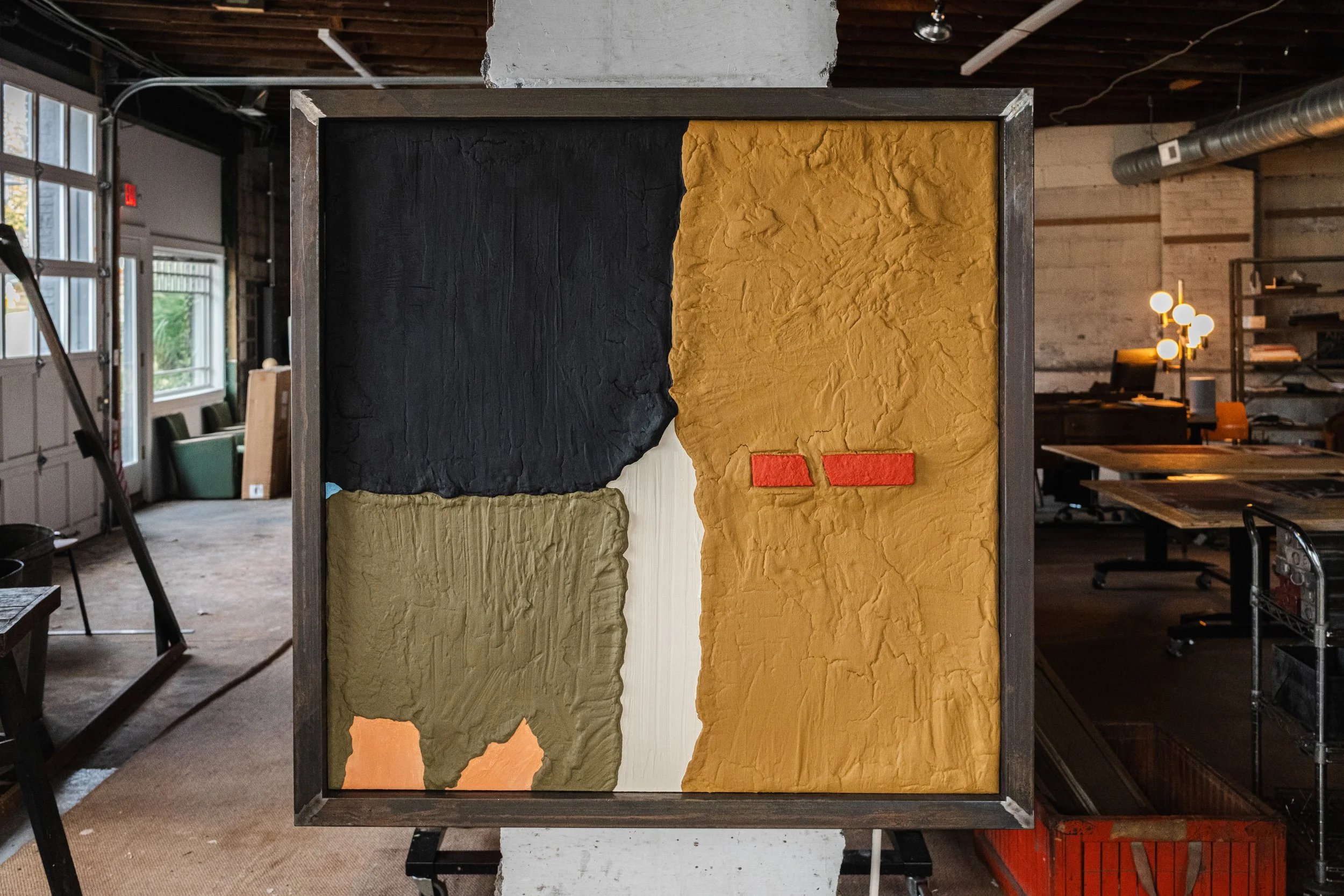 A square colorfield sculpture painting is hanging in the studio. Black, cream, red, pink, blue, ochre.
