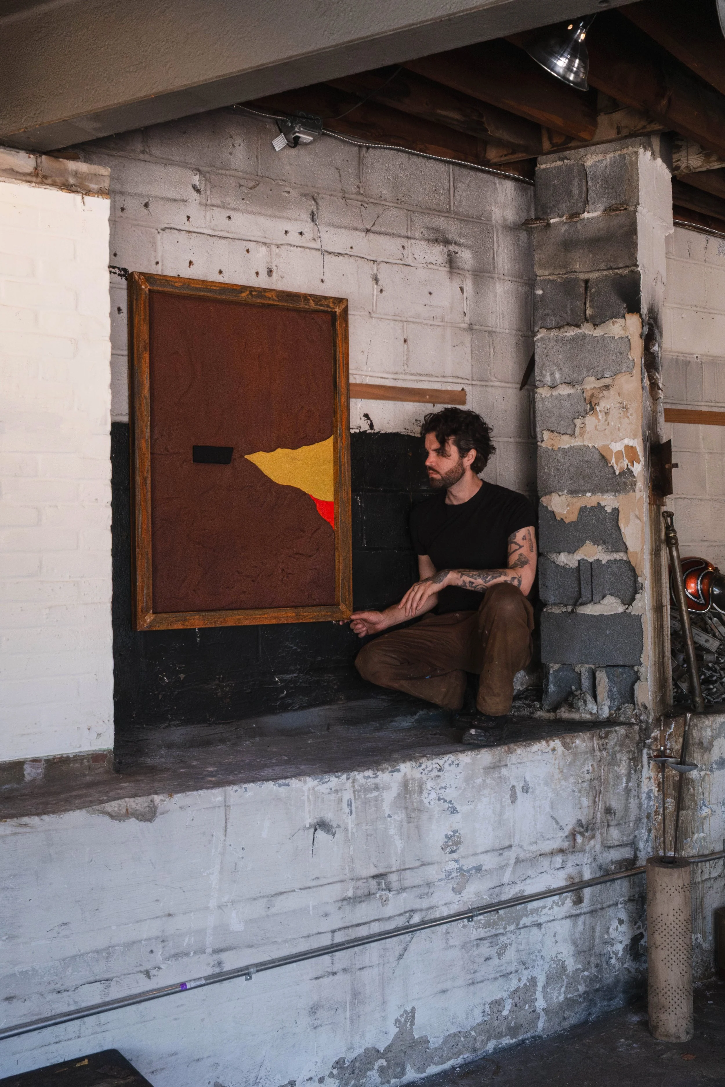 The artist adjusting a framed sculpture painting on the wall of his studio. Industrial space. Rust and scarlet and black and gold.