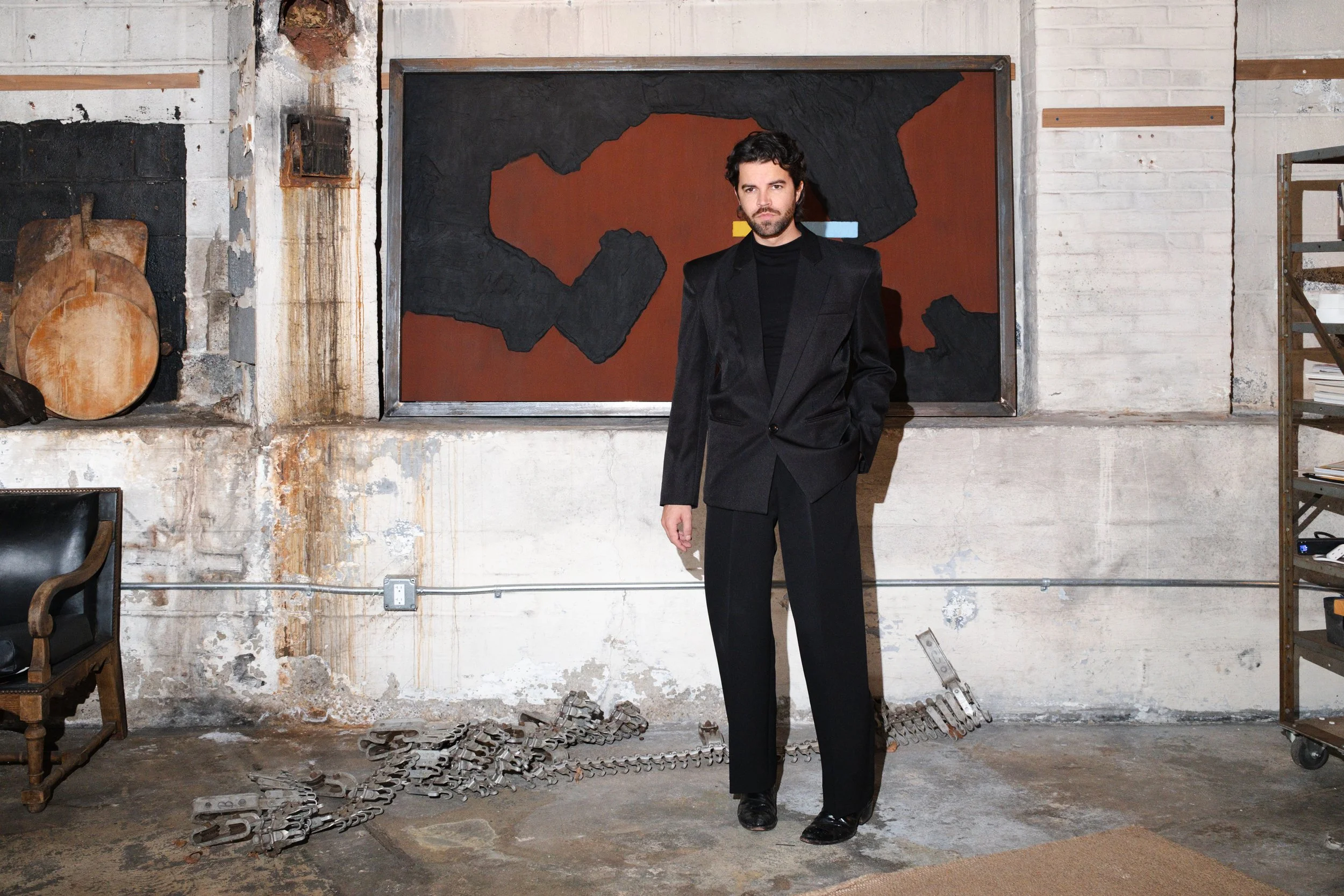 The artist stands in front of a large painting in a black suit.