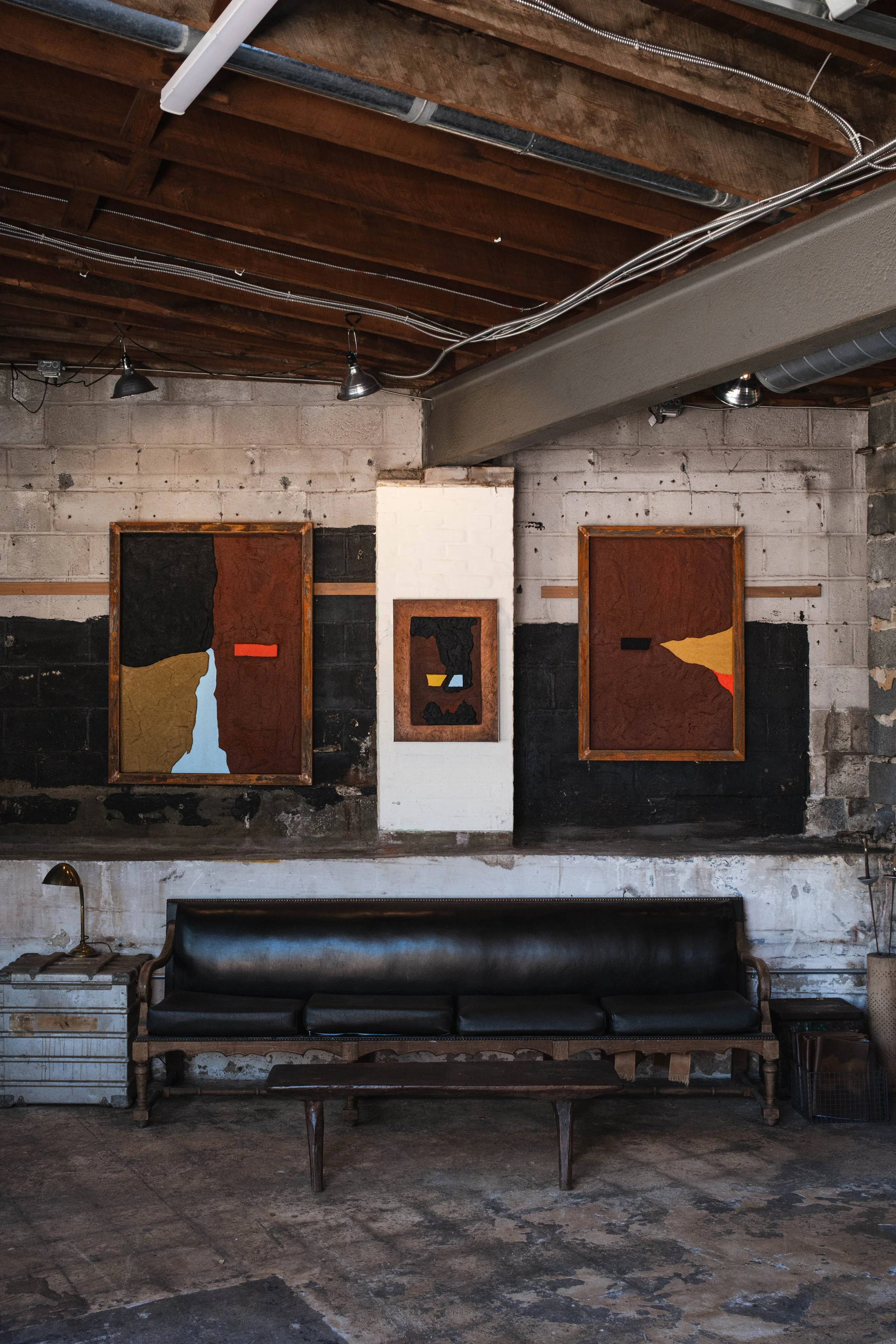 Three color field scultped paintings framed and hanging on the wall of an industrial studio. A black and wood couch sits beneath them.