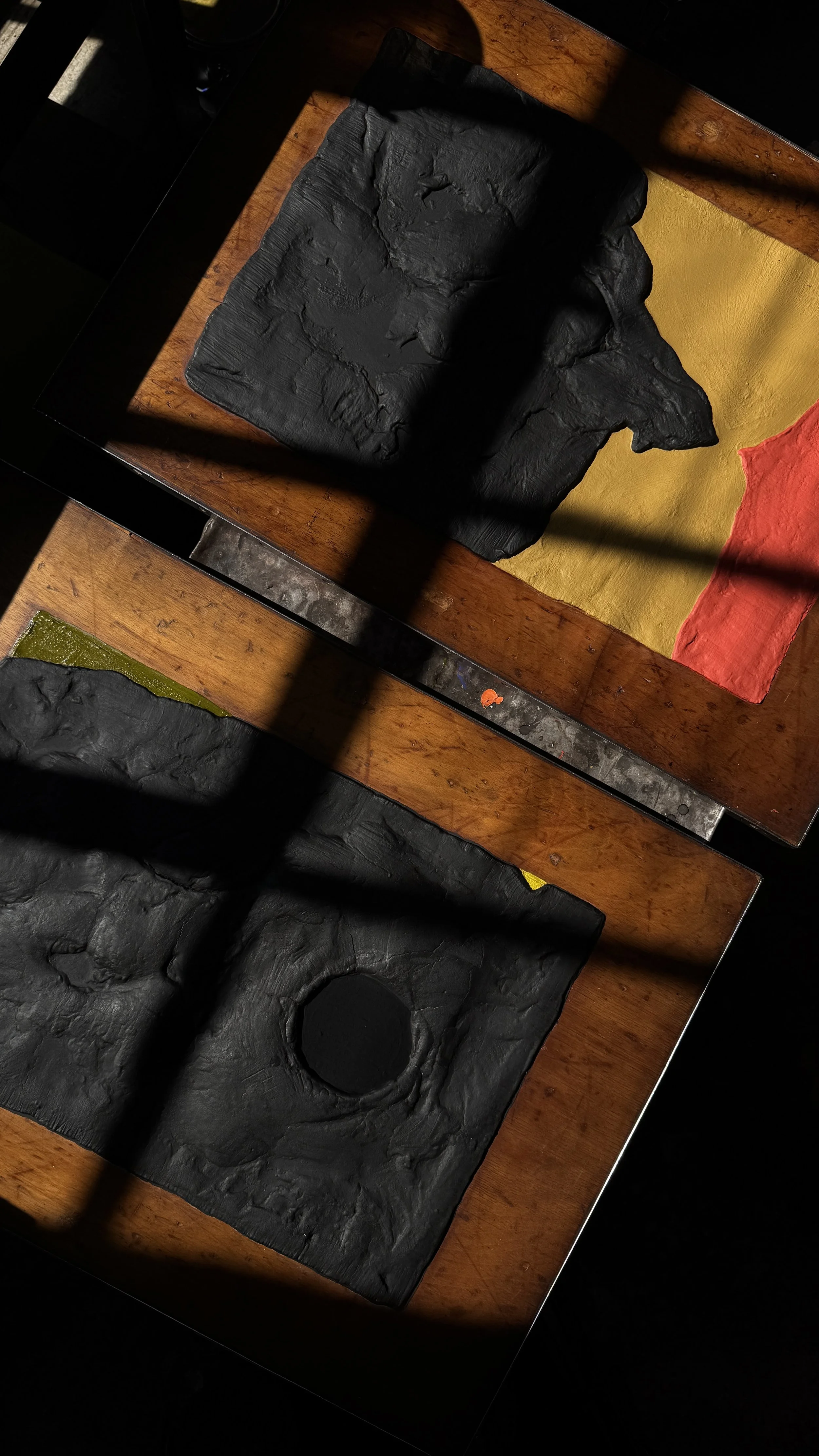 Two paintings in extreme sunlight and shadow being shown side by side on a table, close up, both in black with featured colors.