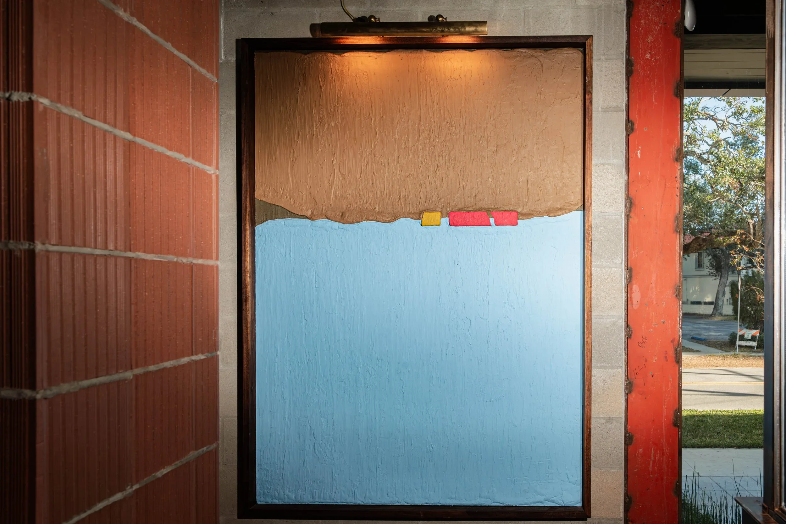 A large colorfield sculpture painting on a CMU block wall next to a window. Blue and tan and red and yellow.