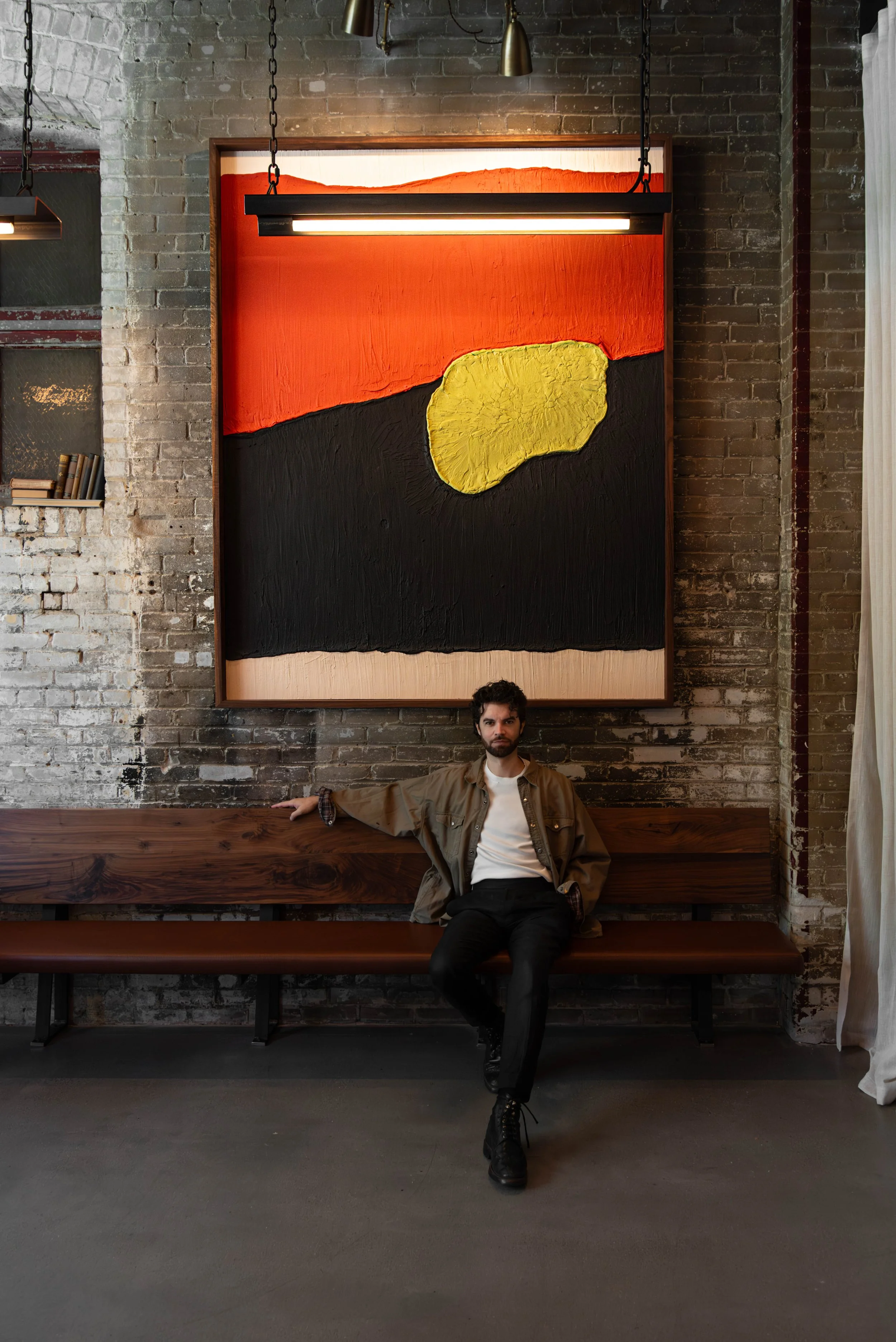 A man sits on a wooden bench against a brick wall with a large abstract painting featuring bold black, red, yellow, and beige colors hanging behind him.