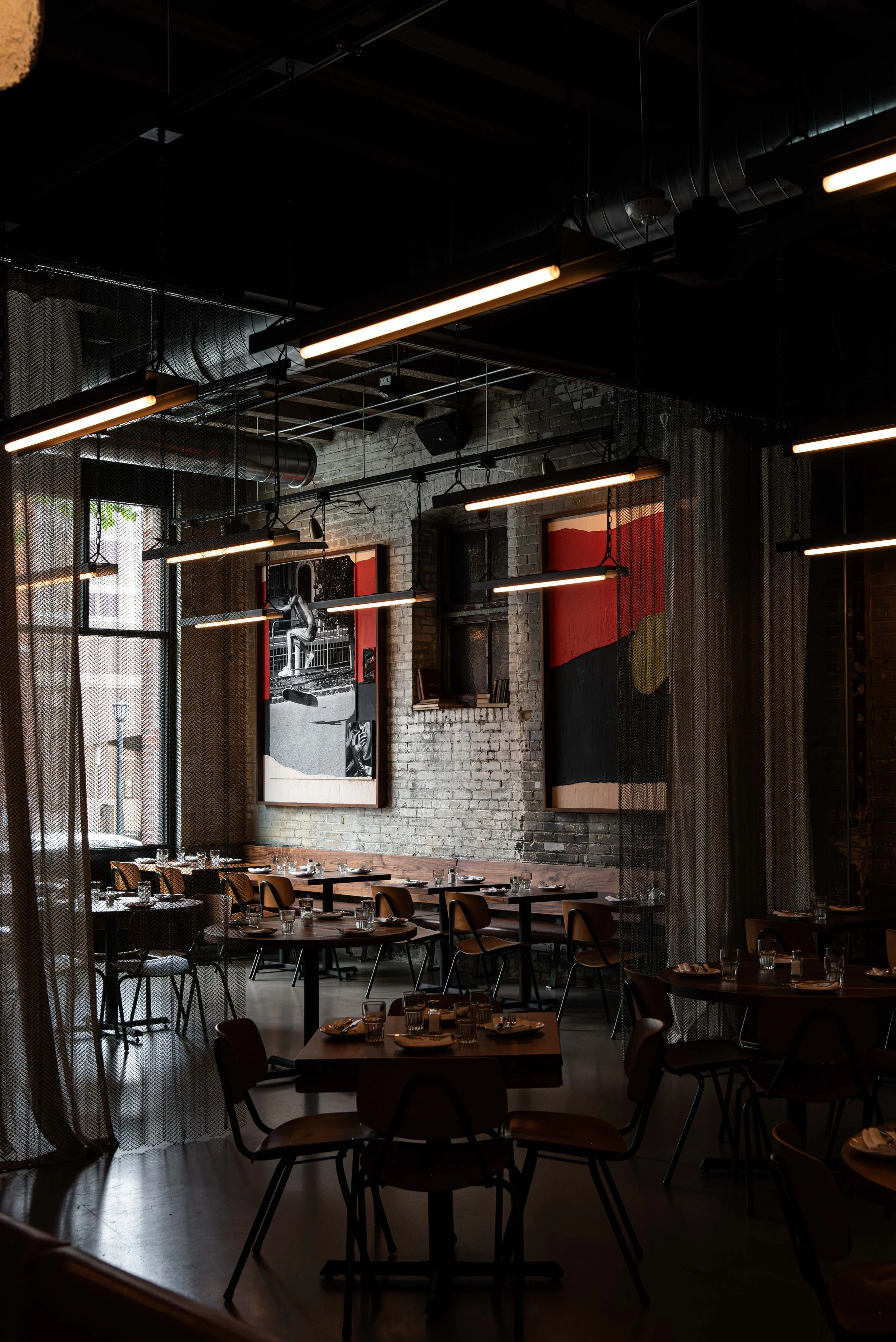 Dimly lit modern restaurant with artwork on exposed brick wall overlooking tables set with glasses and utensils.