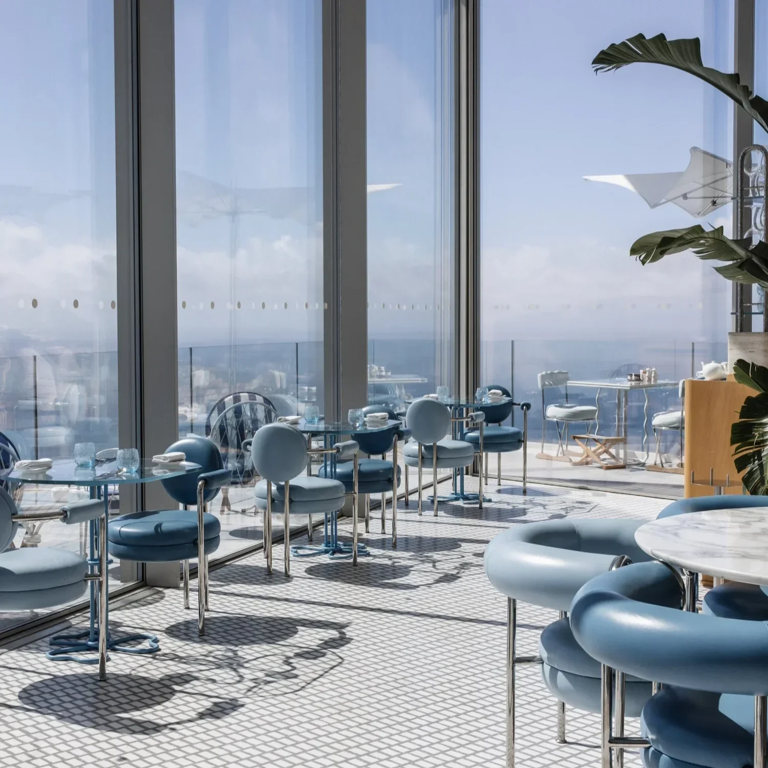A modern restaurant with large glass windows overlooking the ocean, featuring blue and white chairs, marble tables, and a patterned tiled floor with natural sunlight.