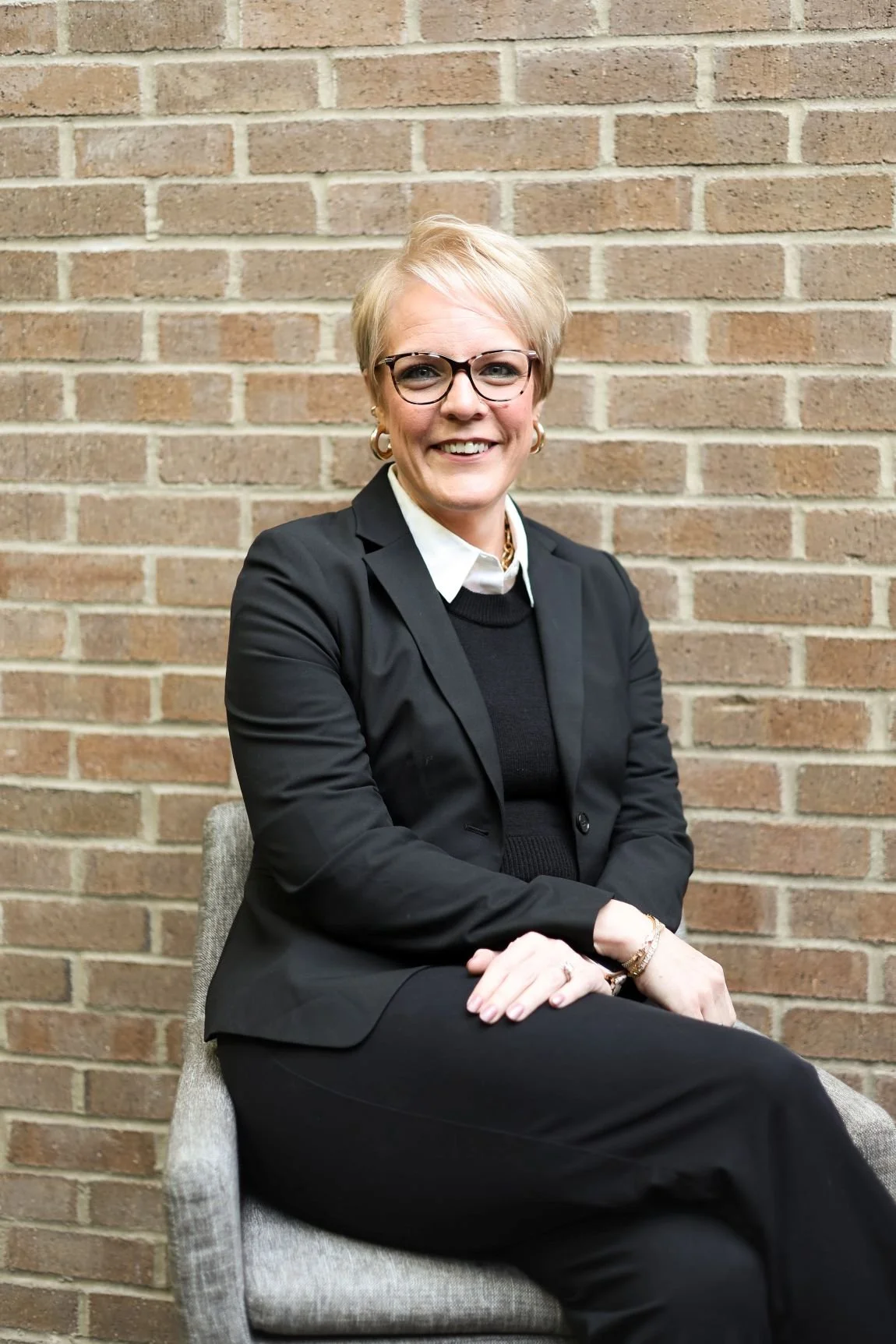 Mary Hollst in black suit white blouse sitting in front of brick wall smiling