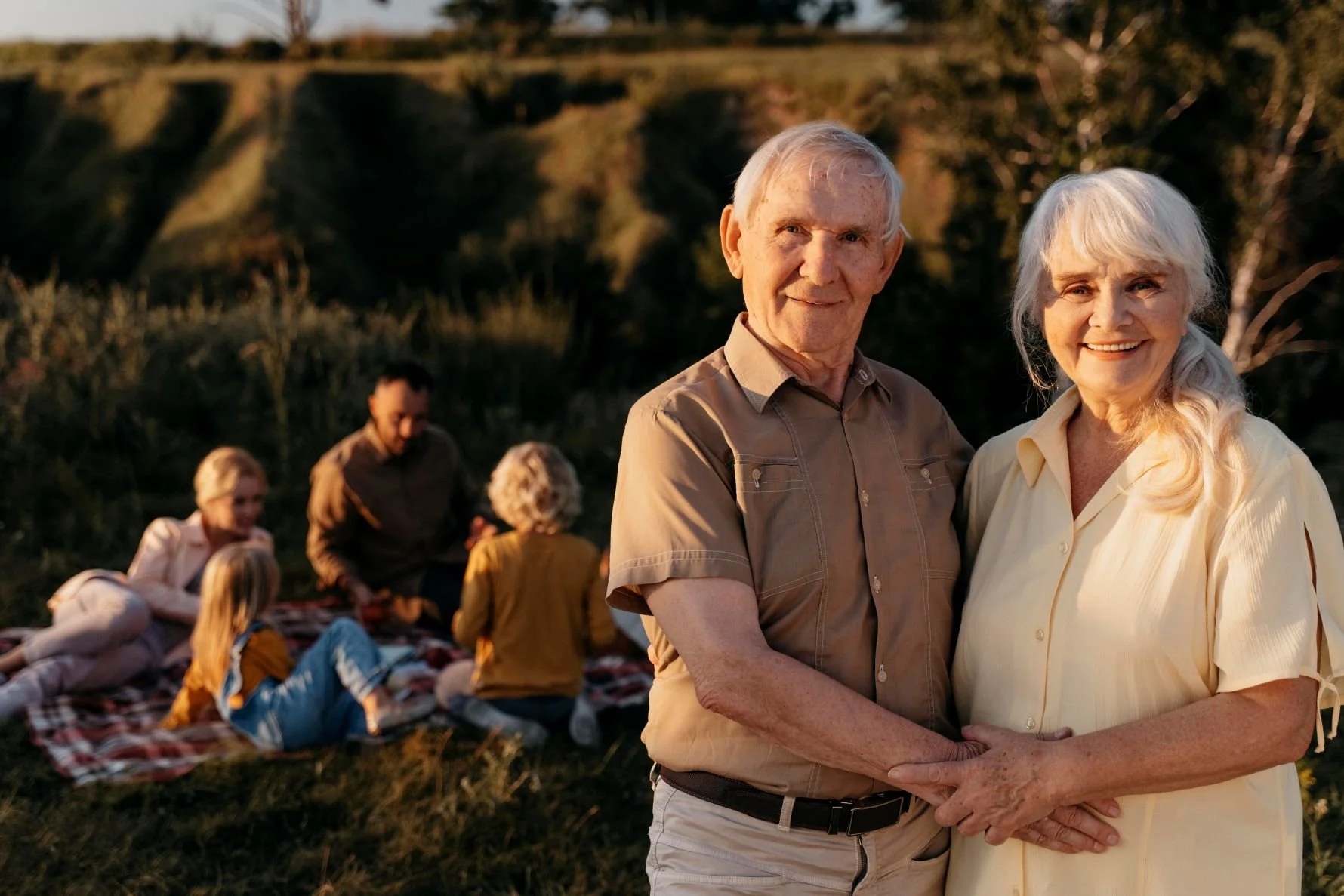elderly couple holding hands in foreground with family having a picnic in the background