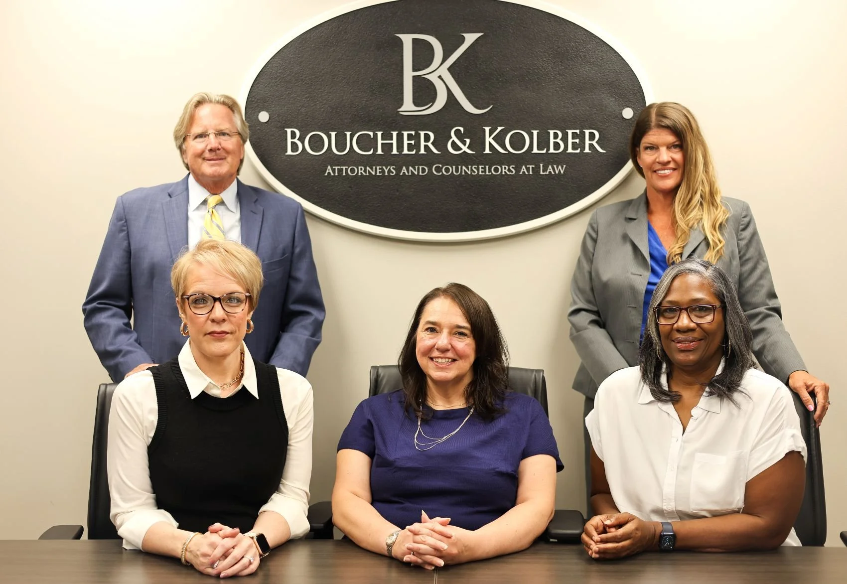 Five members of the boucher and kolber legal team sitting at a desk with firm's sign on wall