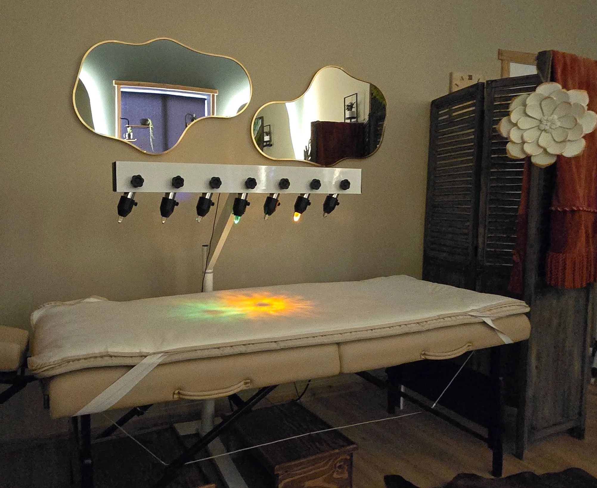 Bedroom vanity area with two gold-edged mirrors, a wall-mounted light fixture, a padded bench, and a black folding room divider with a white flower decoration.