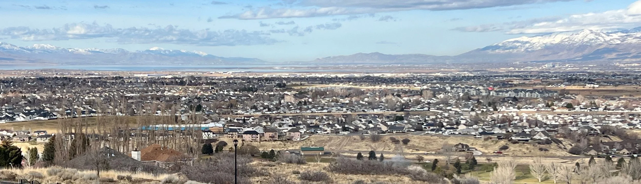 View of a town called Spanish Fork with many houses, fields, and some trees. Mountain range with snow-capped peaks in the background, under a mostly cloudy sky.