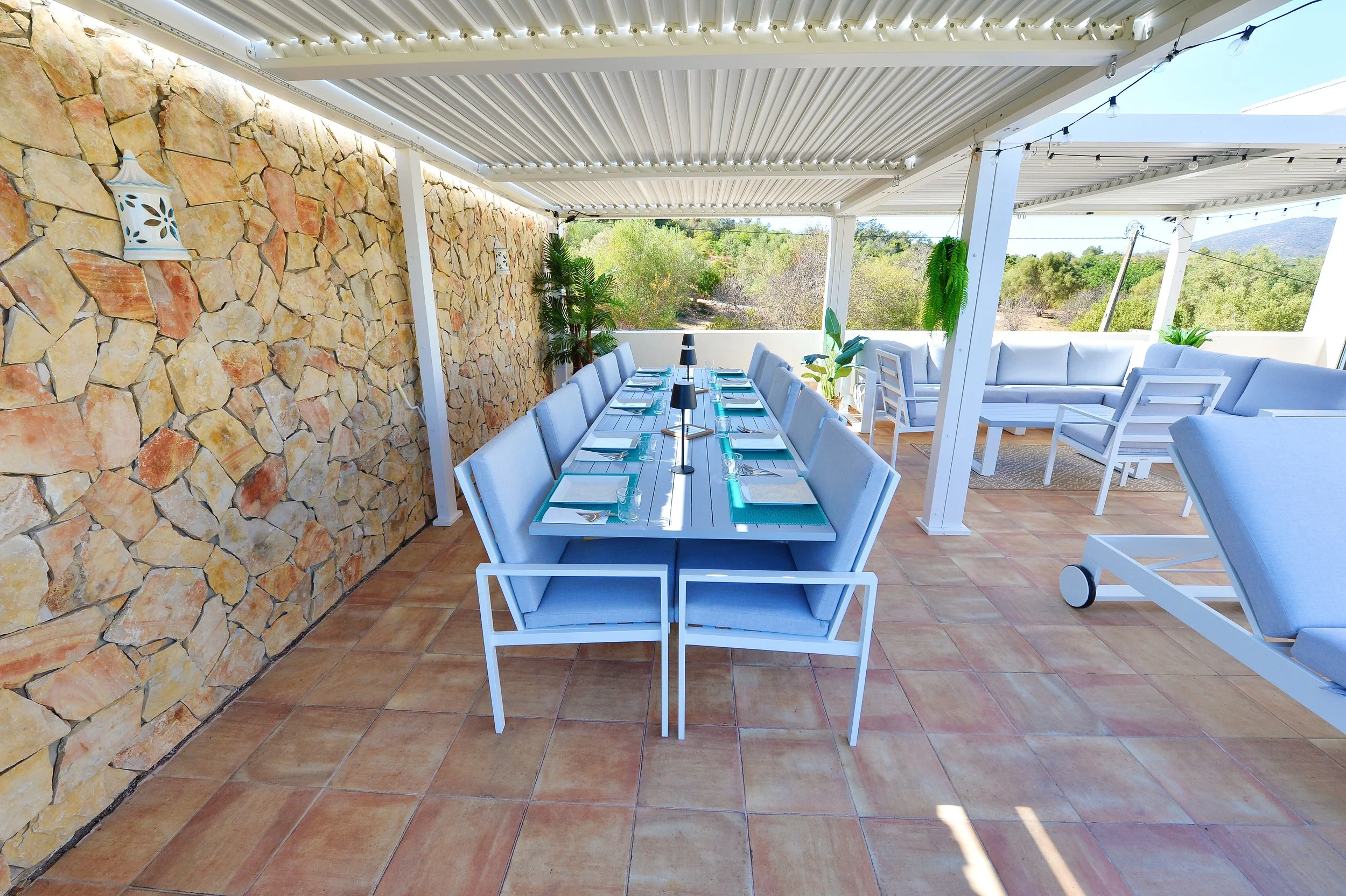 Outdoor patio with a long dining table set with plates, forks, knives, napkins, and candle centerpieces. The patio has terracotta tile flooring, a stone wall with lanterns, potted plants, and seating areas with cushioned chairs and sofas. The patio i