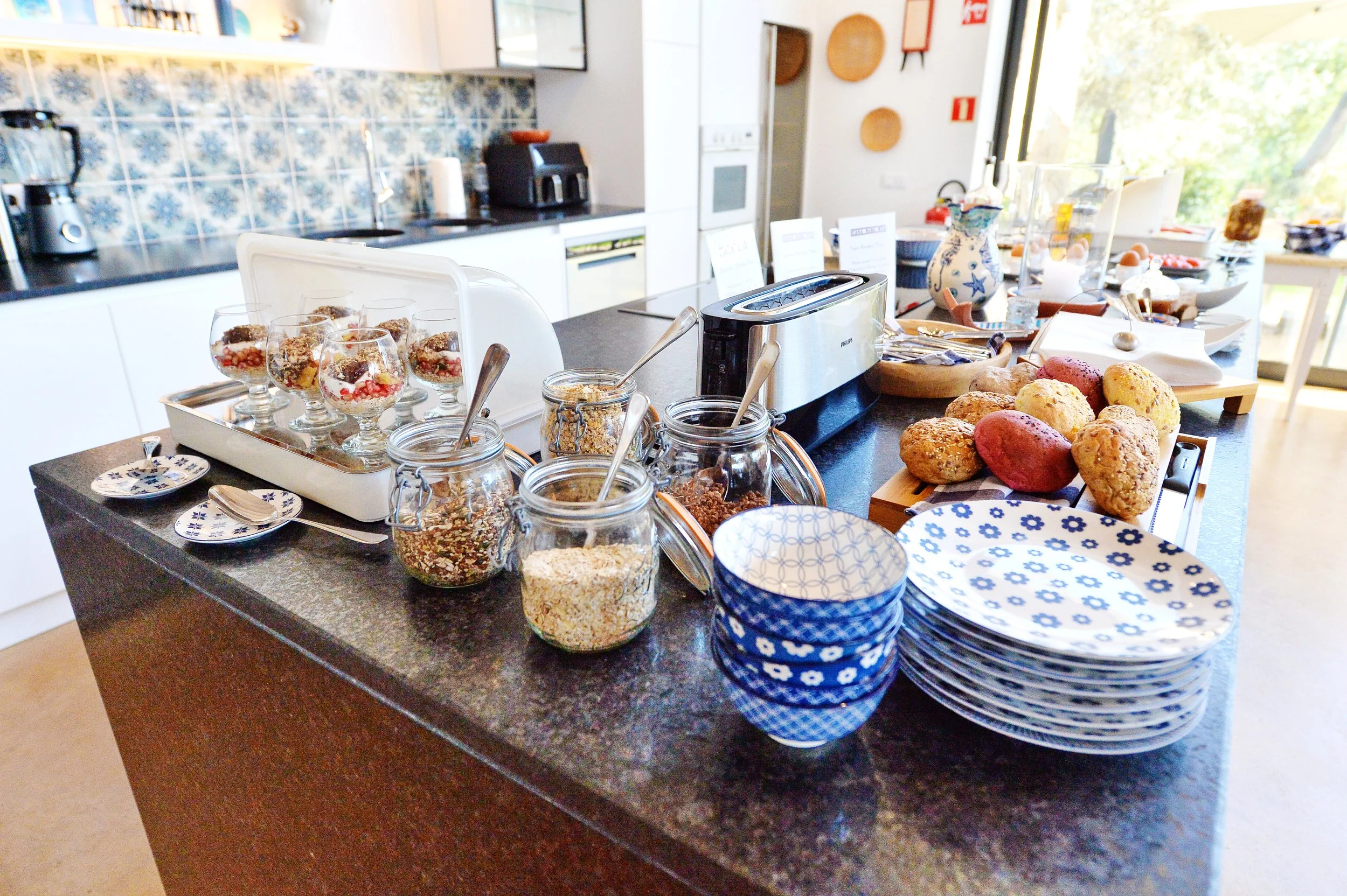 A kitchen counter set up for a breakfast buffet with bowls of fruit, jars of granola, plates, and bread rolls, with kitchen appliances and decorative items in the background.