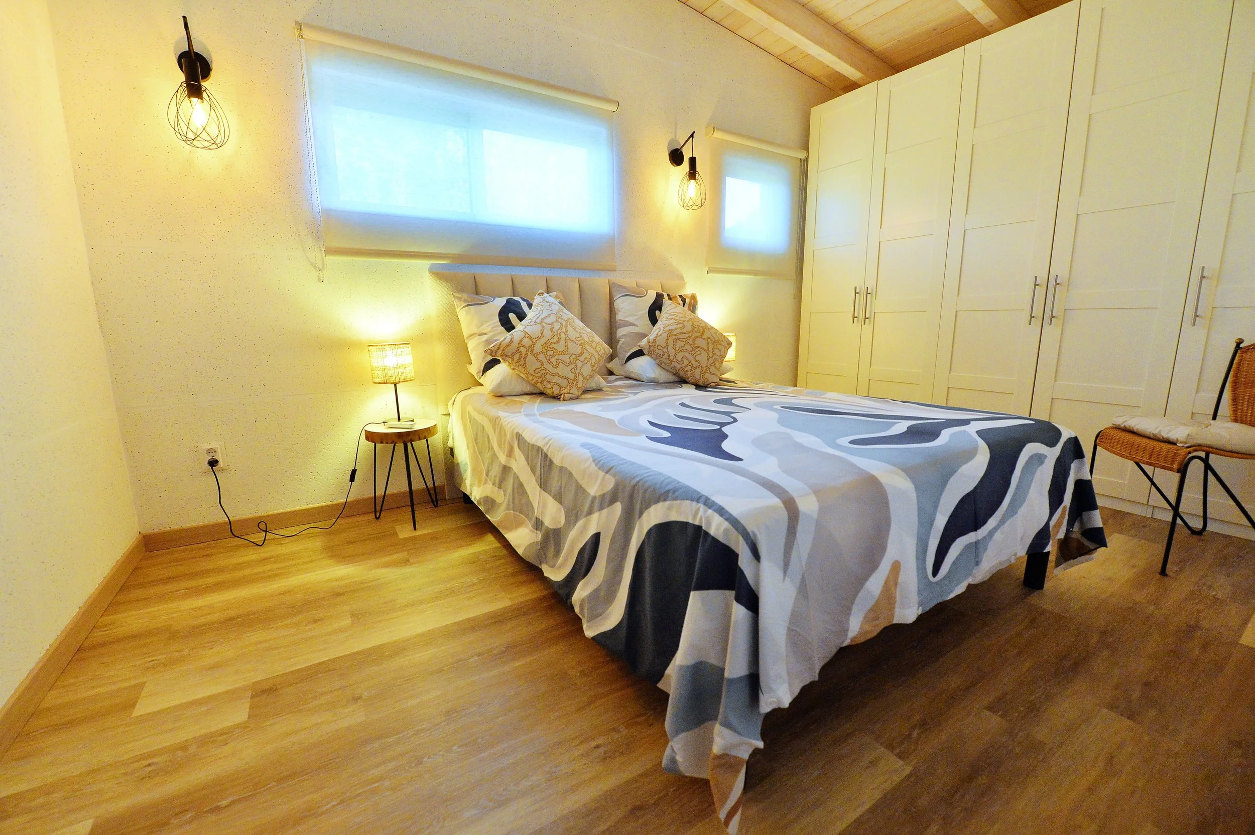A cozy bedroom featuring a bed with patterned bedding and decorative pillows, a small side table with a lamp, window blinds, built-in white wardrobe, wooden flooring, and warm lighting from wall lamps.