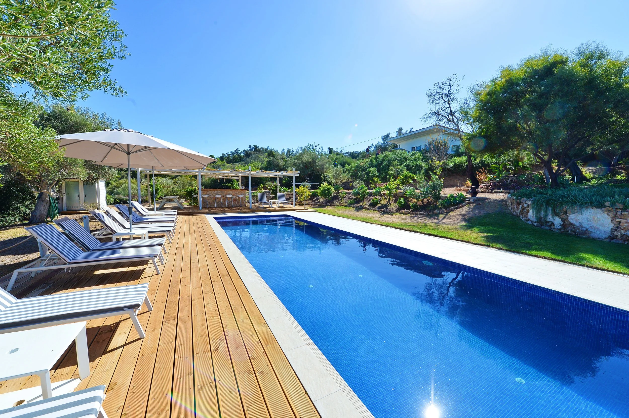 Swimming pool surrounded by lounge chairs with striped cushions and umbrellas in a sunny outdoor setting with trees and a house in the background.