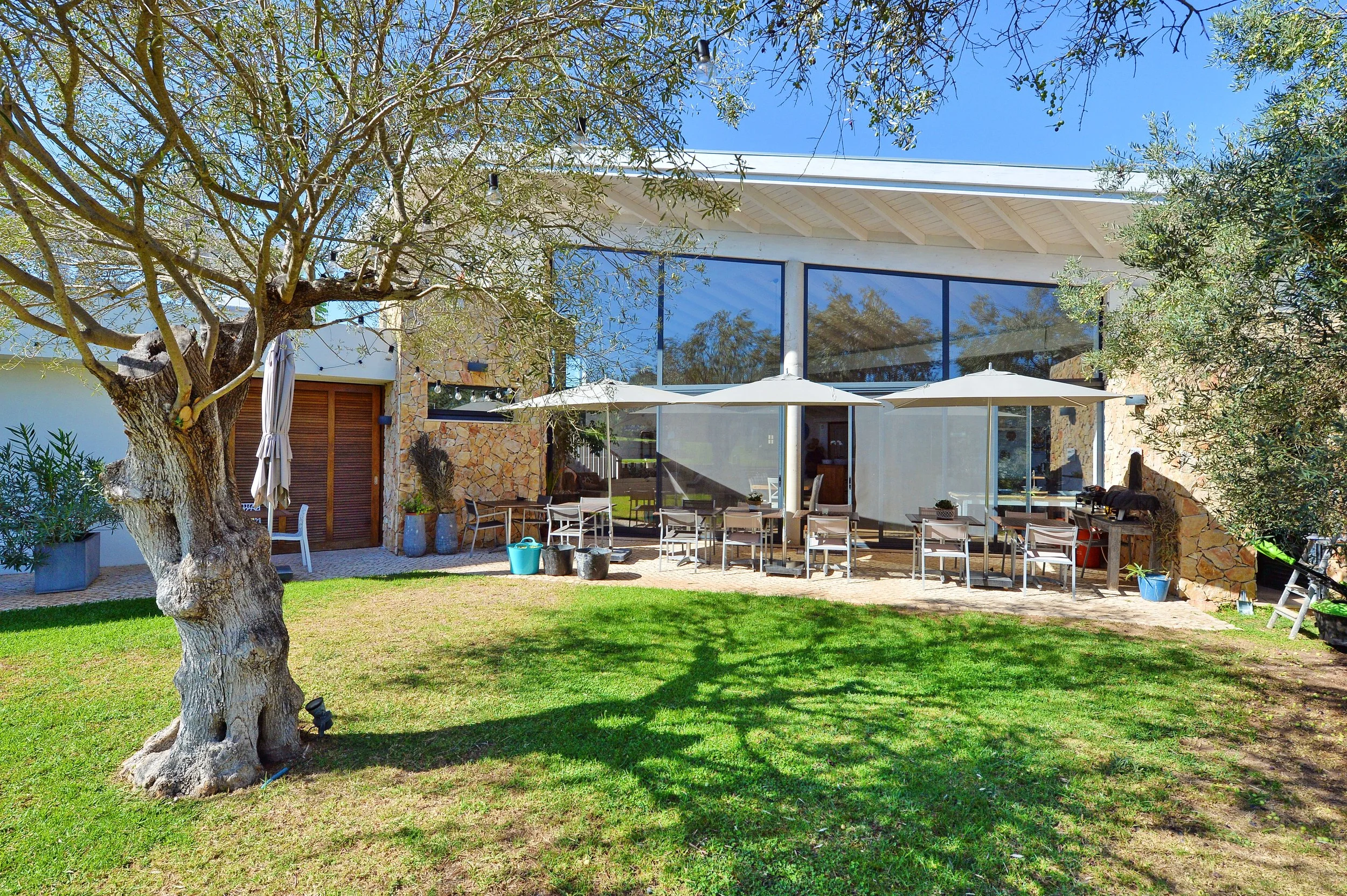 Modern house with large glass windows, outdoor patio with tables and umbrellas, surrounded by trees and green grass.