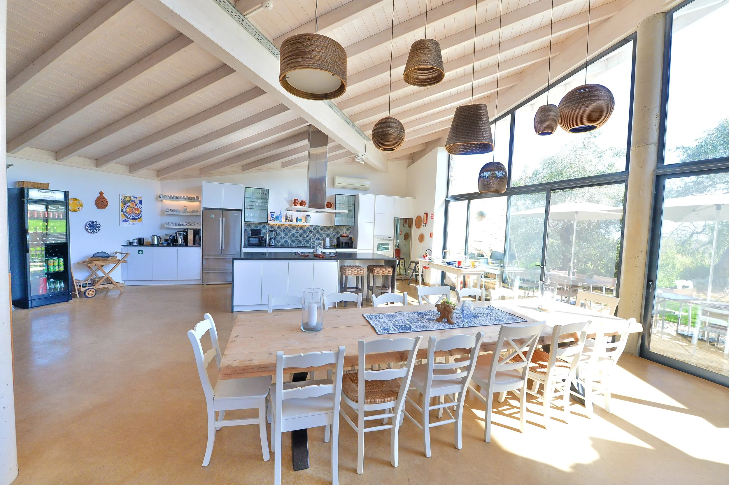 Open-concept kitchen and dining area with white cabinets, a large wooden dining table, white chairs, and large windows letting in natural light.