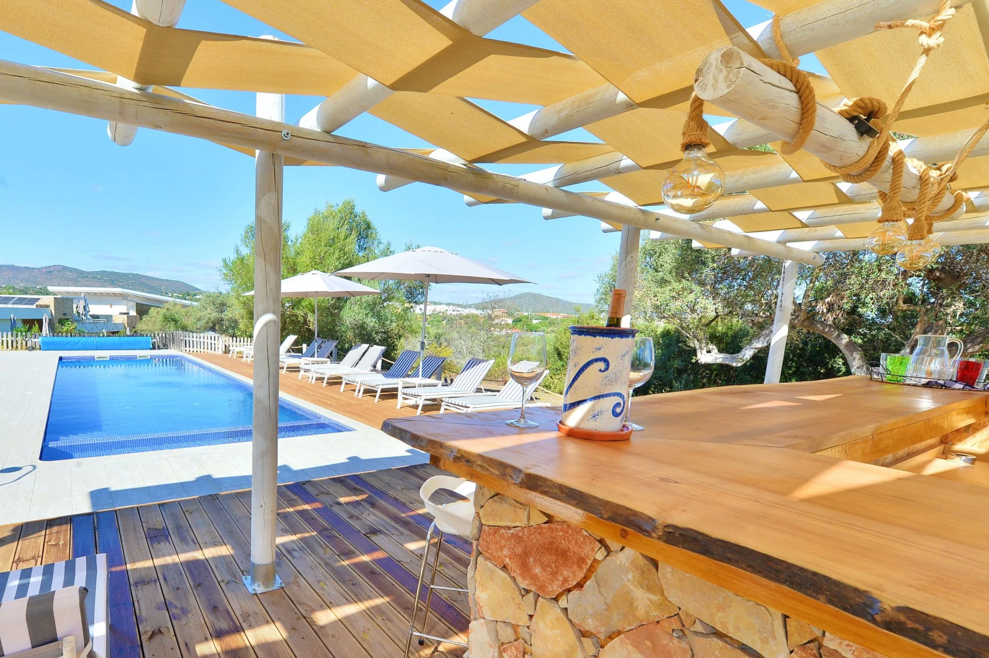 Outdoor swimming pool with lounge chairs and umbrellas, wooden deck, bar area with wood countertop, glassware, and decorative lighting, lush greenery, and mountainous landscape in the background on a sunny day.