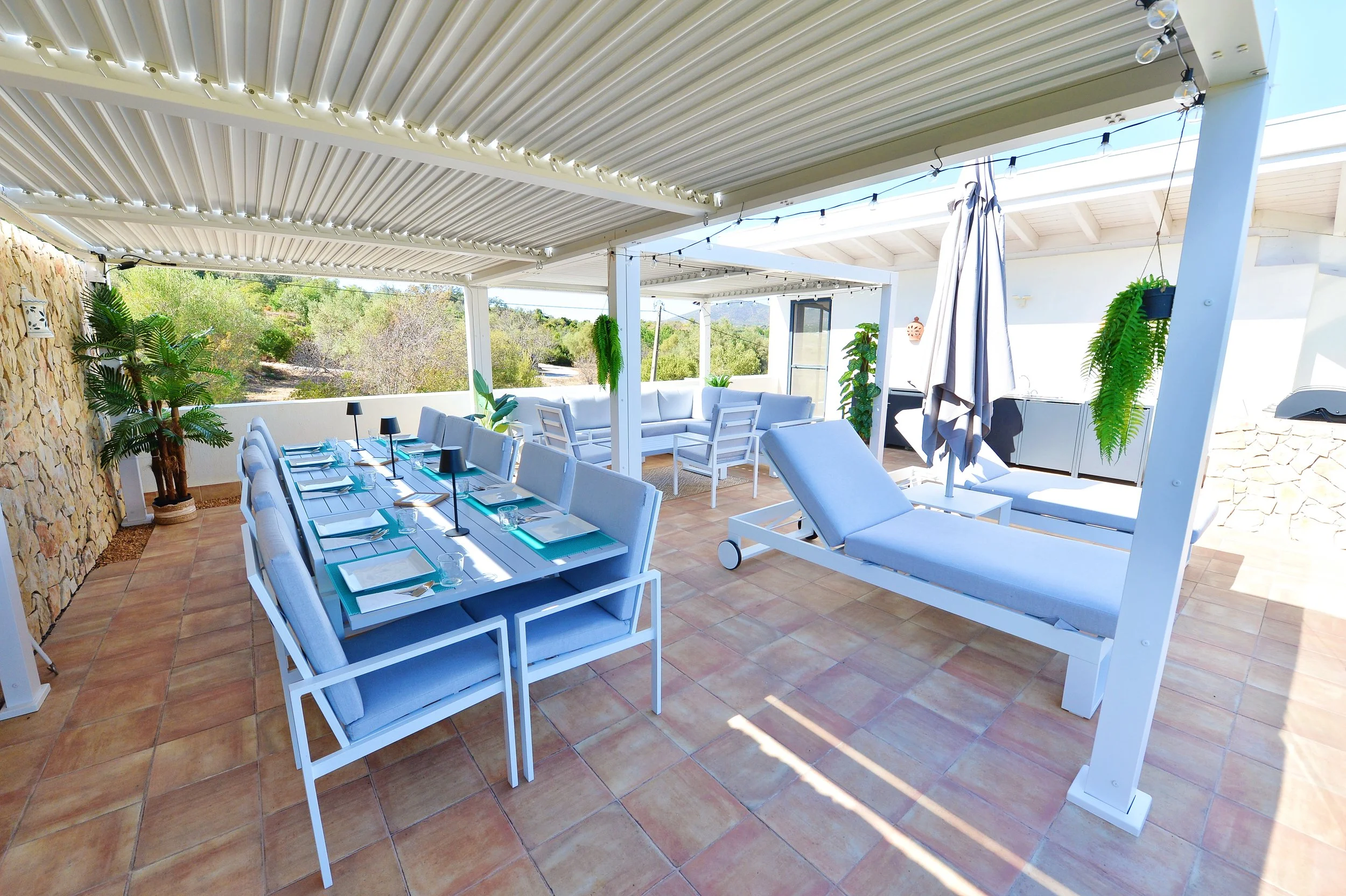 Outdoor patio with a dining table set for a meal, lounge chairs, and hanging plants under a corrugated metal roof, overlooking nature.