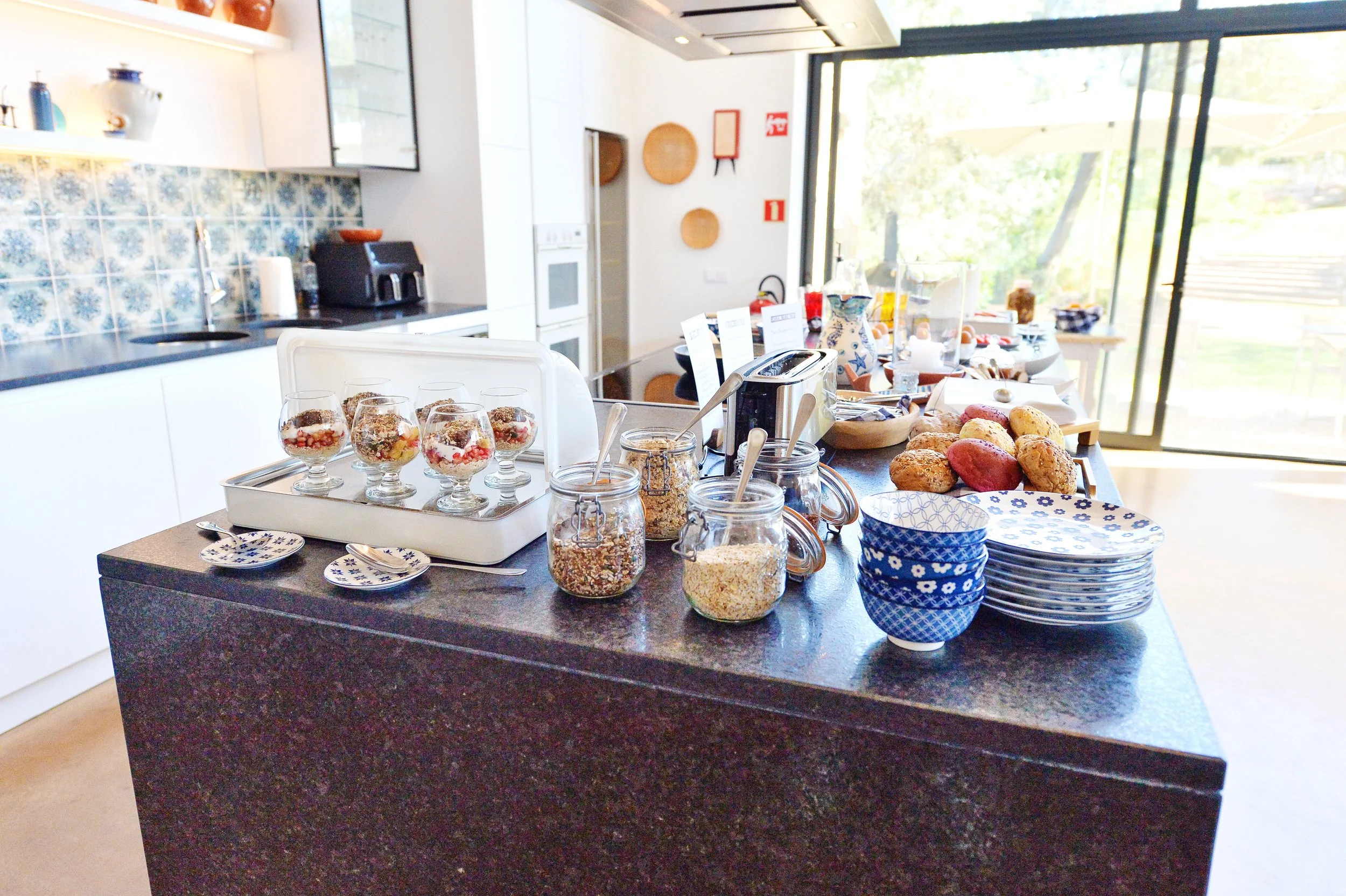 A breakfast or buffet setup on a kitchen island, featuring glasses of fruit parfaits, bowls, jars of oats and granola, plates, and baked goods near a large sliding glass door with natural light.