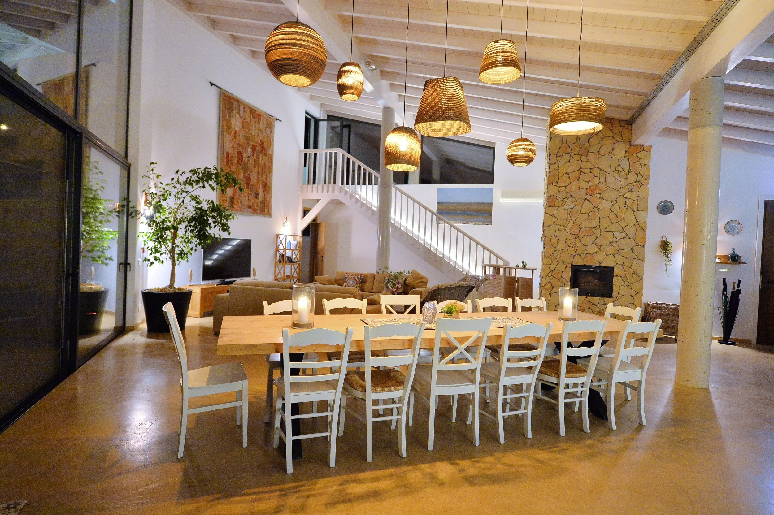 Open-concept living and dining area with wooden floors, white walls, and a high ceiling. A large wooden dining table with white chairs is in the foreground, decorated with candles. Behind, a cozy living space features a beige couch, a television, and