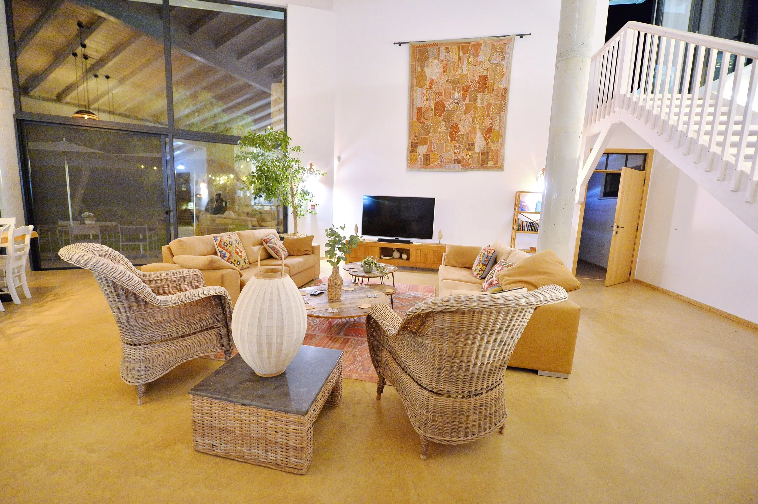 A cozy living room with a large glass window, beige sofas, wicker chairs, a round coffee table, potted plants, wall art, a television, and a staircase leading upstairs.