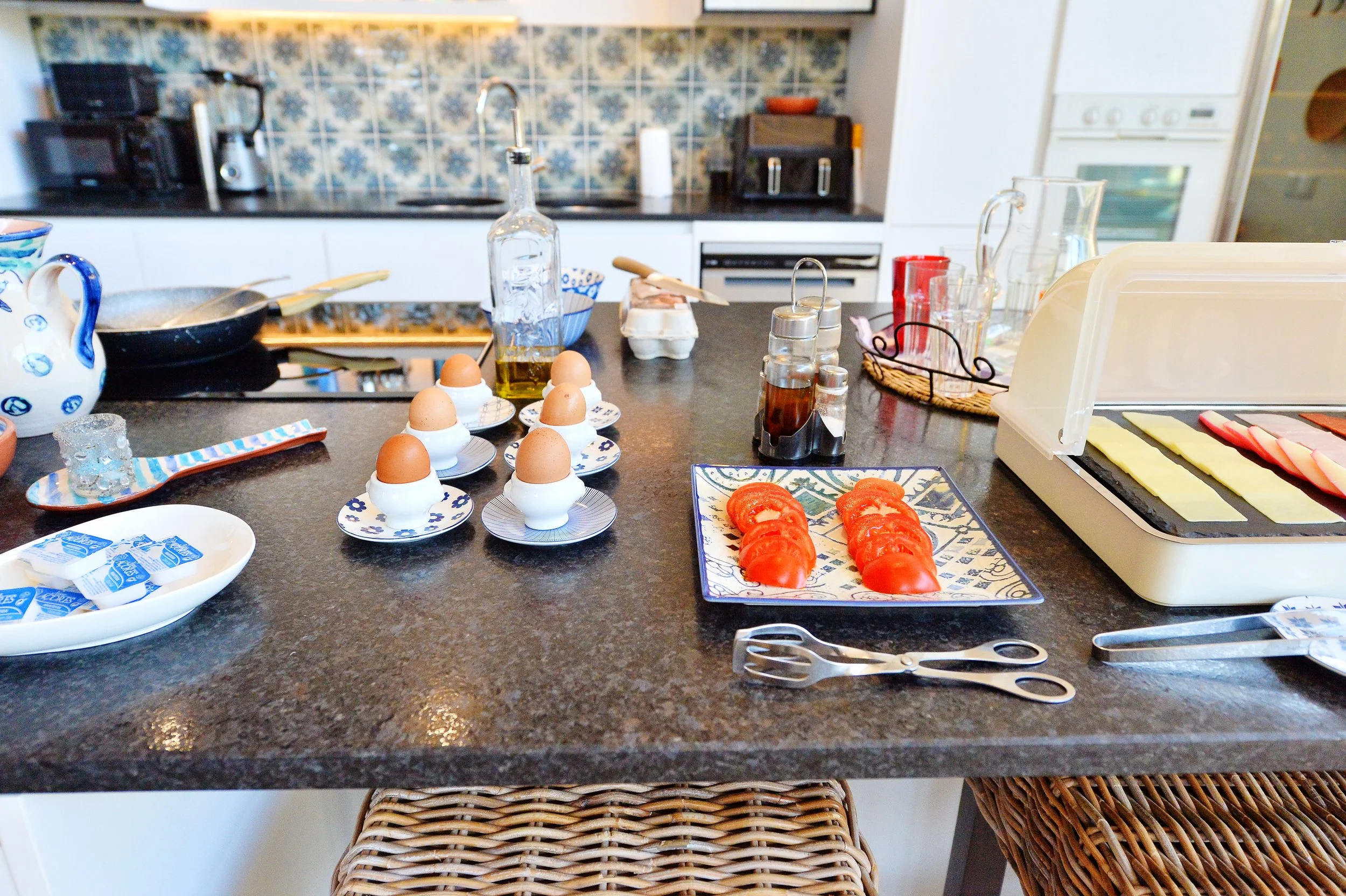 Kitchen countertop with eggs in egg cups, sliced tomatoes, condiments, cheese slices, and utensils, with a stove and cabinets in the background.